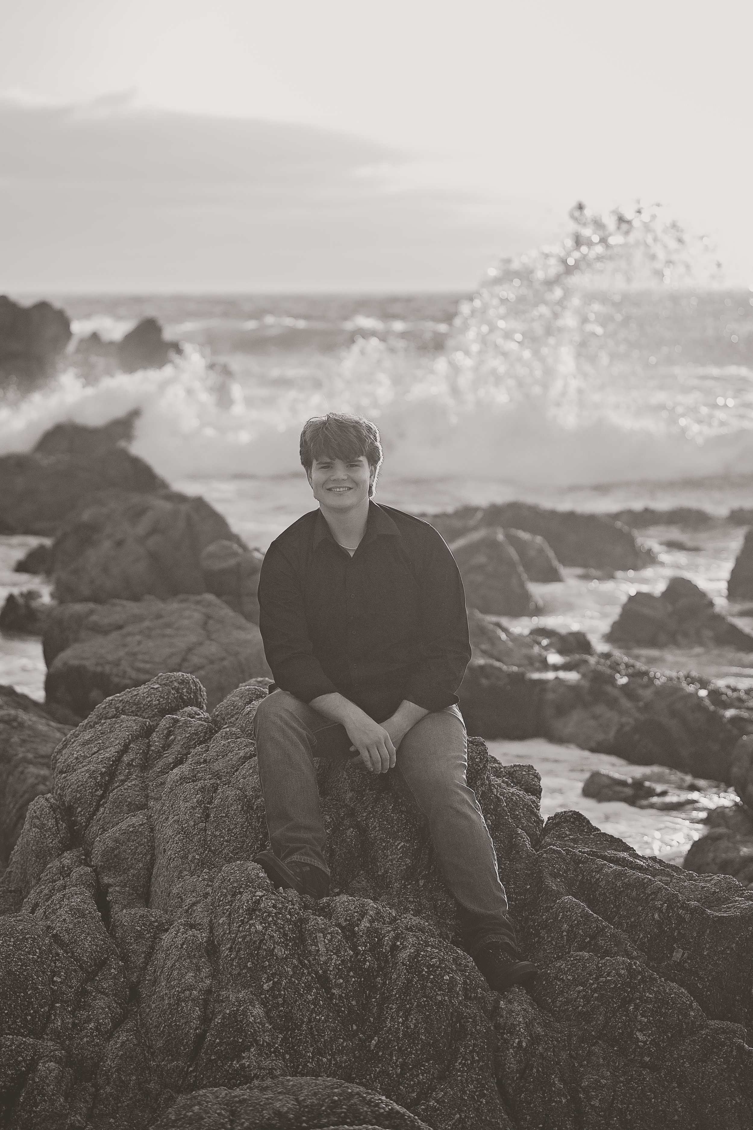Black and white senior portrait of Gianni sitting on the rocks as waves crash along the Asilomar coastline.