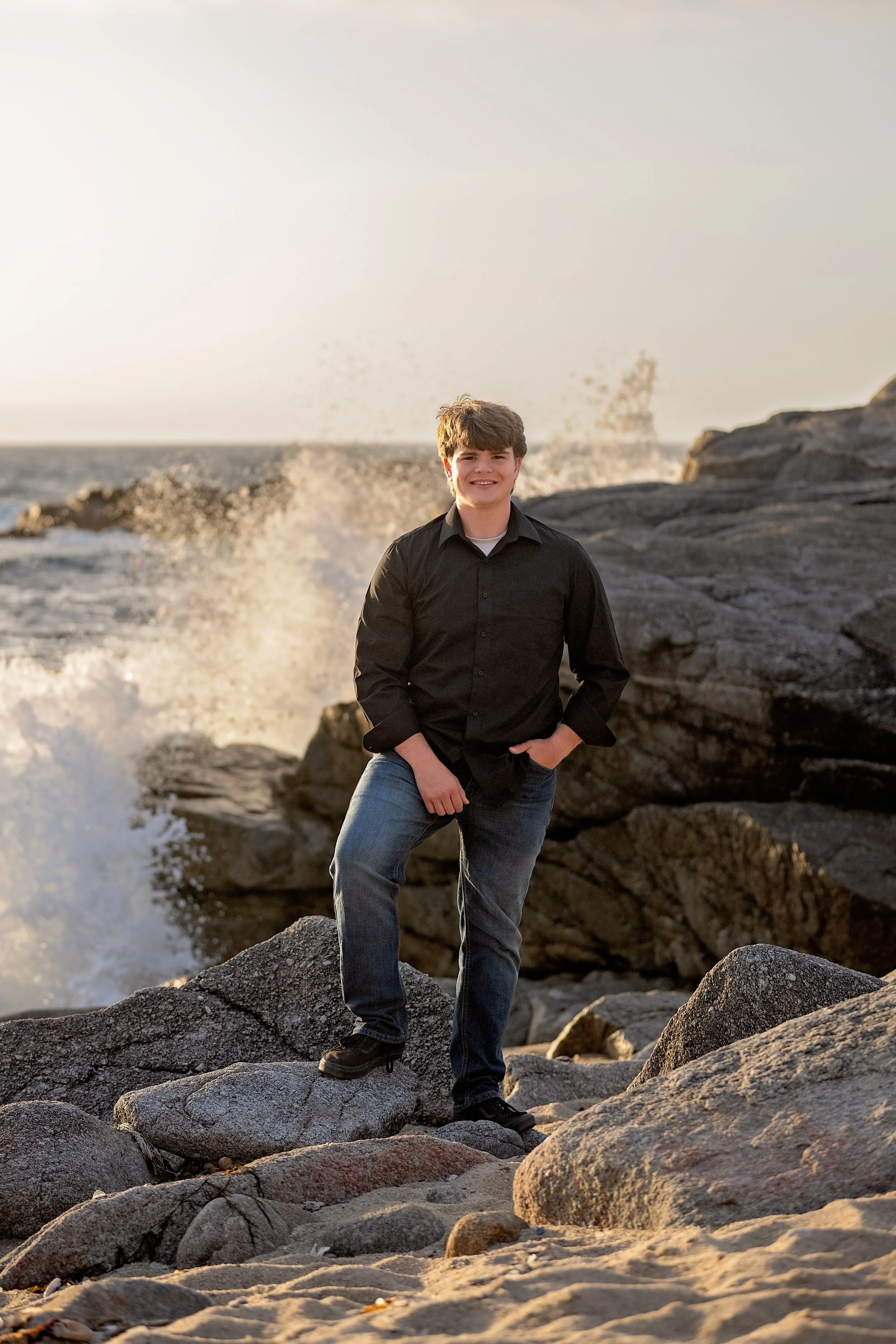 Gianni standing on coastal rocks with a crashing wave behind him during sunset in Pacific Grove.