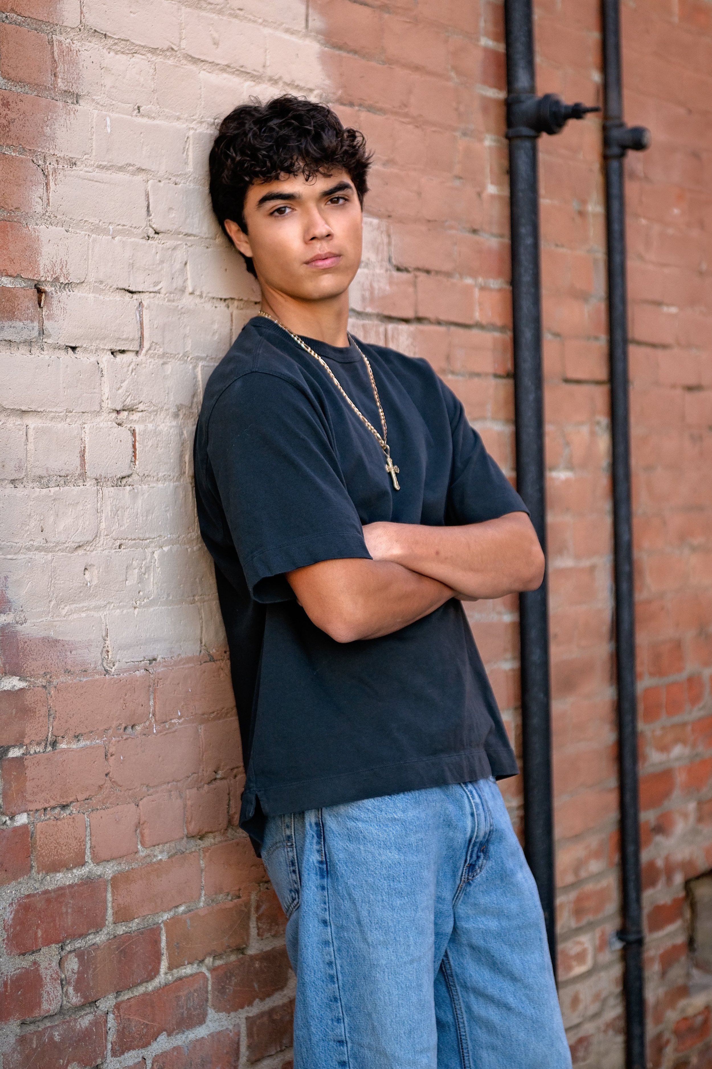 Senior portrait of Cruz leaning against a brick wall in Downtown Salinas wearing a black shirt and jeans.