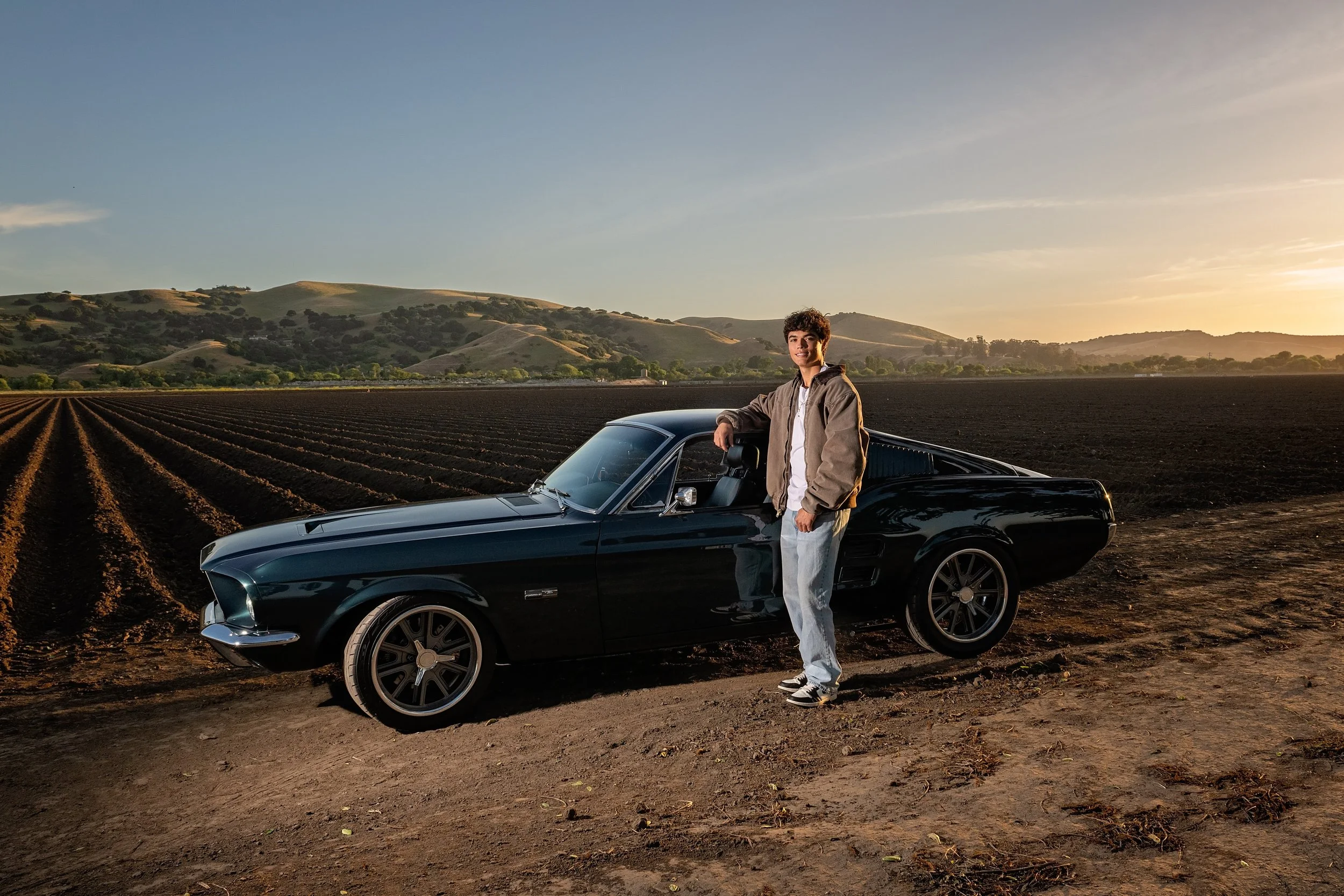 Cruz leaning against his vintage Ford Mustang with Salinas Valley farmland and hills behind him at sunset.