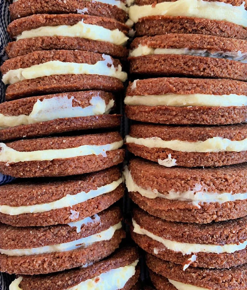 Homemade low carb chocolate sandwich cookies with cream cheese filling stacked on a white plate, crispy chocolate almond flour cookies with white cream showing
