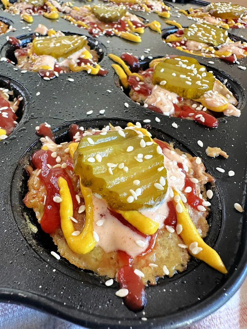 High protein cheeseburger muffins with a cheesy center in a muffin tin next to sugar-free burger sauce and raw vegetables