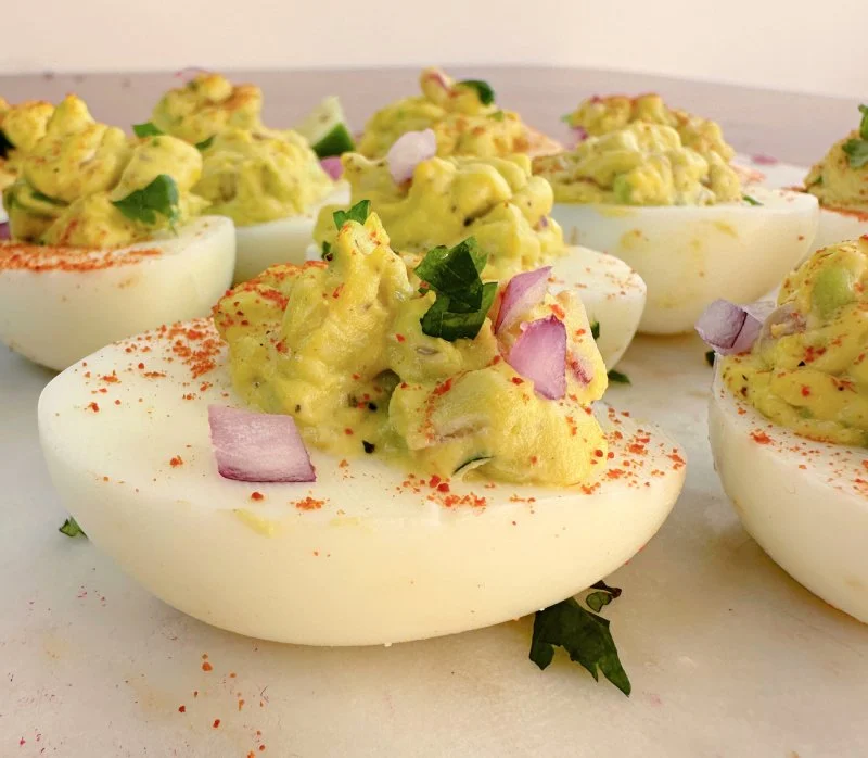 Avocado deviled eggs with green guacamole-style filling, diced avocado pieces visible, finished with paprika on a white platter