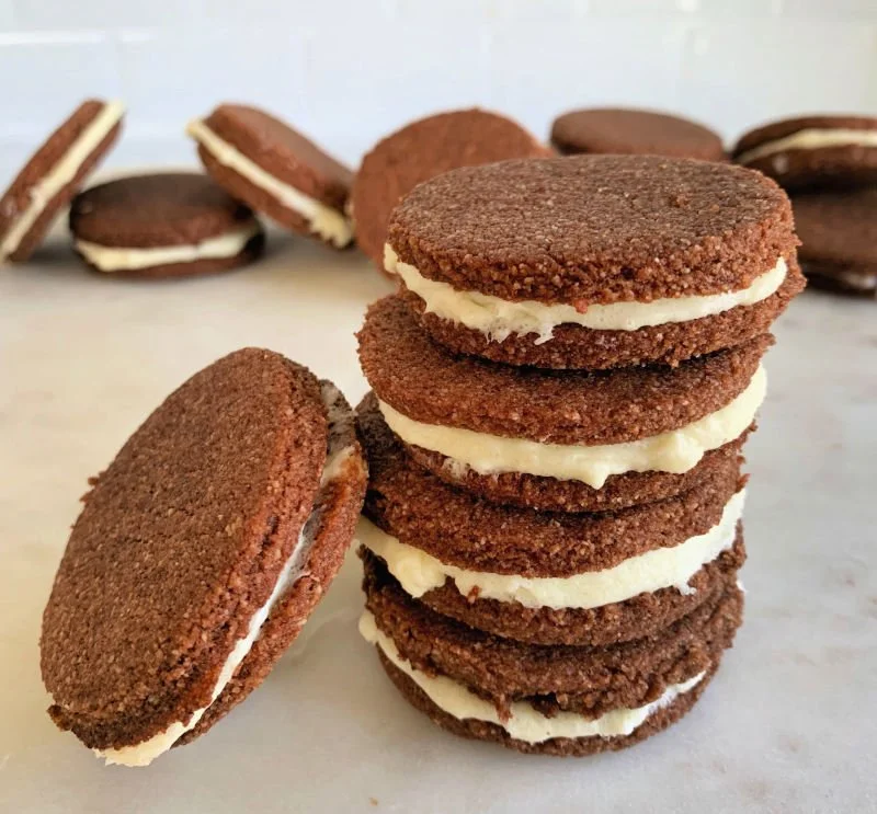 Homemade low carb chocolate sandwich cookies with cream cheese filling stacked on a white plate, crispy chocolate almond flour cookies with white cream showing