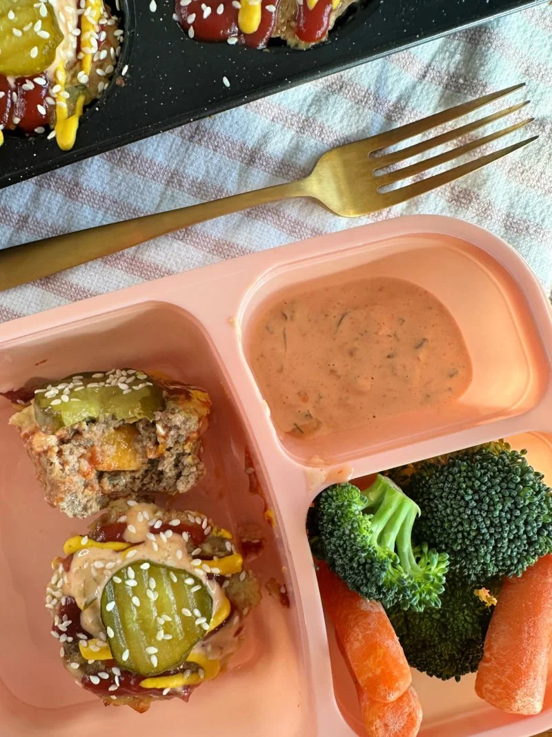 High protein cheeseburger muffins with a cheesy center in a muffin tin next to sugar-free burger sauce and raw vegetables