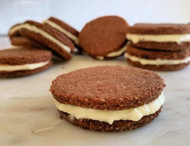 Homemade low carb chocolate sandwich cookies with cream cheese filling stacked on a white plate, crispy chocolate almond flour cookies with white cream showing