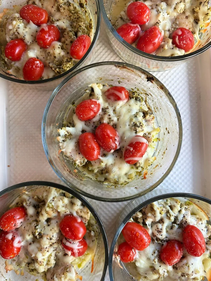 Five creamy pesto chicken meal prep bowls with cherry tomatoes, melted mozzarella, and cauliflower rice in oven-safe glass containers