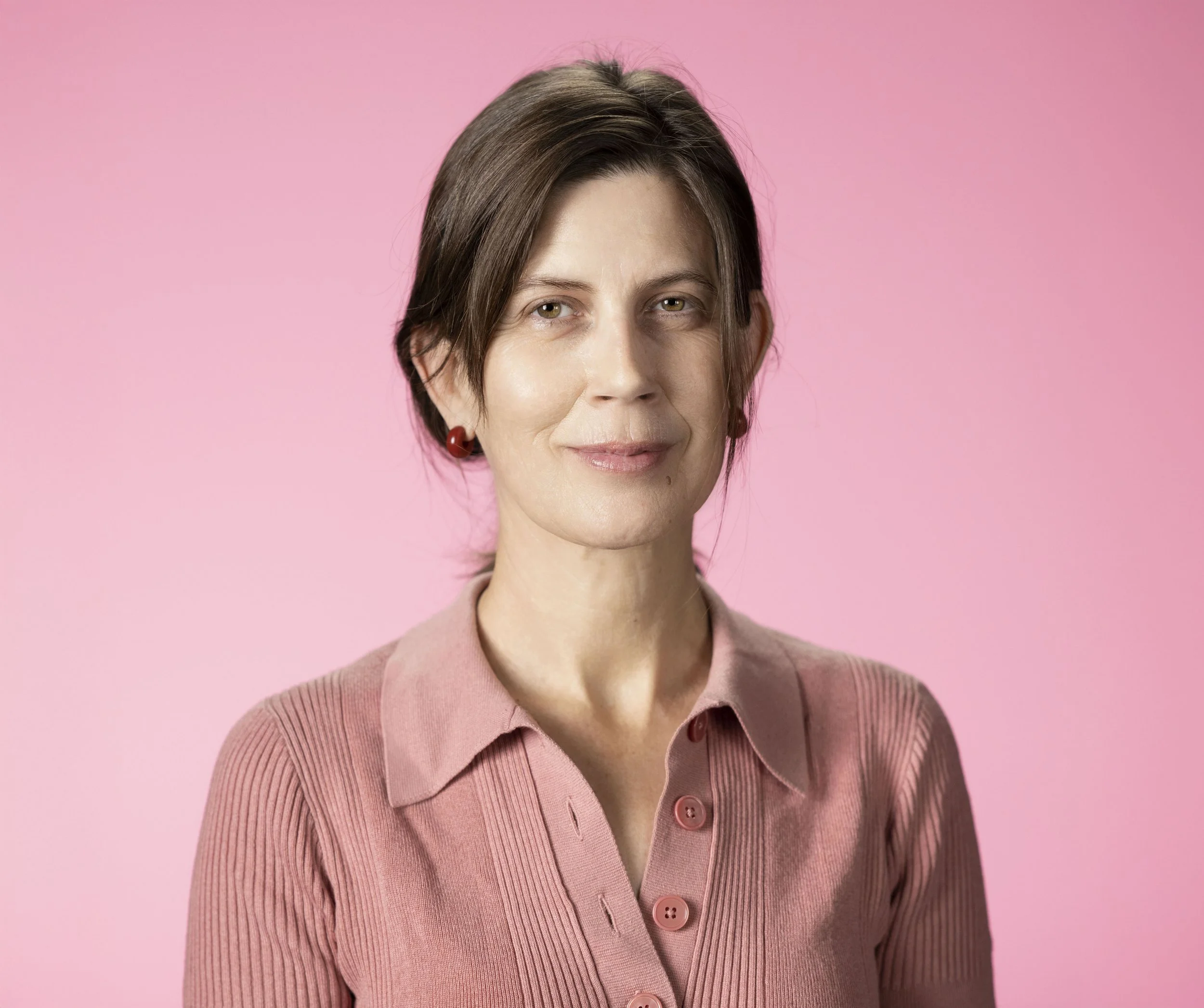 A woman with short brown hair wearing a pink collared shirt and red earrings, standing against a pink background.