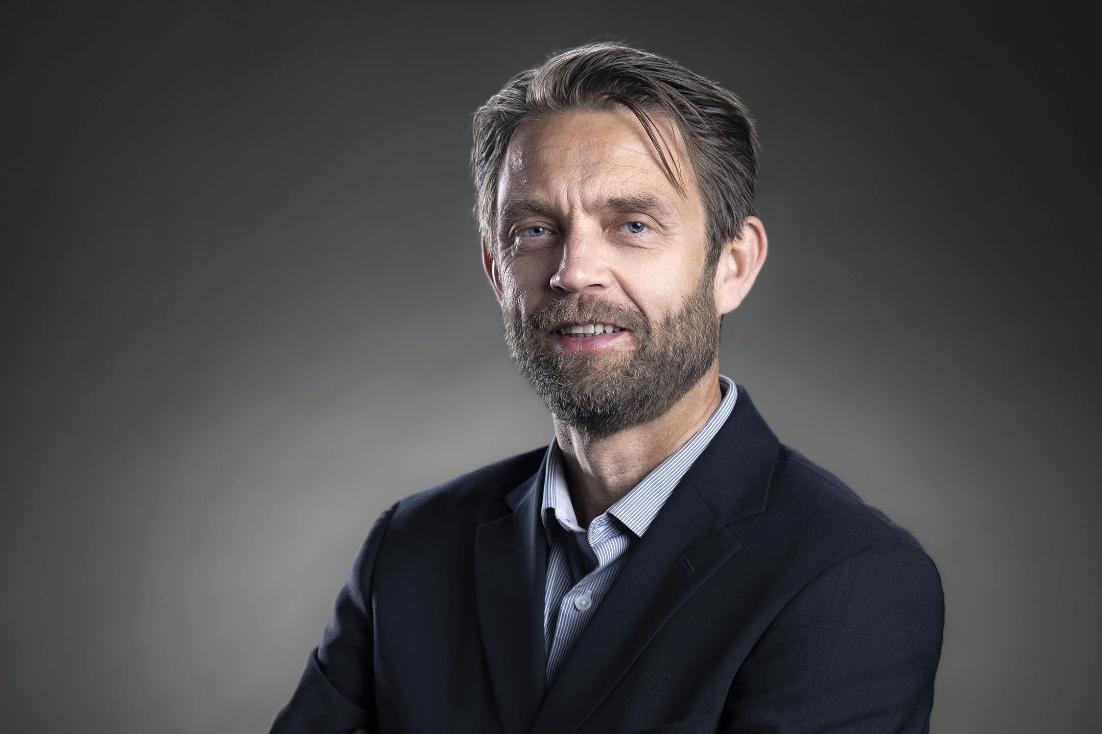 A middle-aged man with light skin, blue eyes, a beard, and brown hair with some gray, wearing a dark suit and a collared shirt, looking at the camera against a gray gradient background.