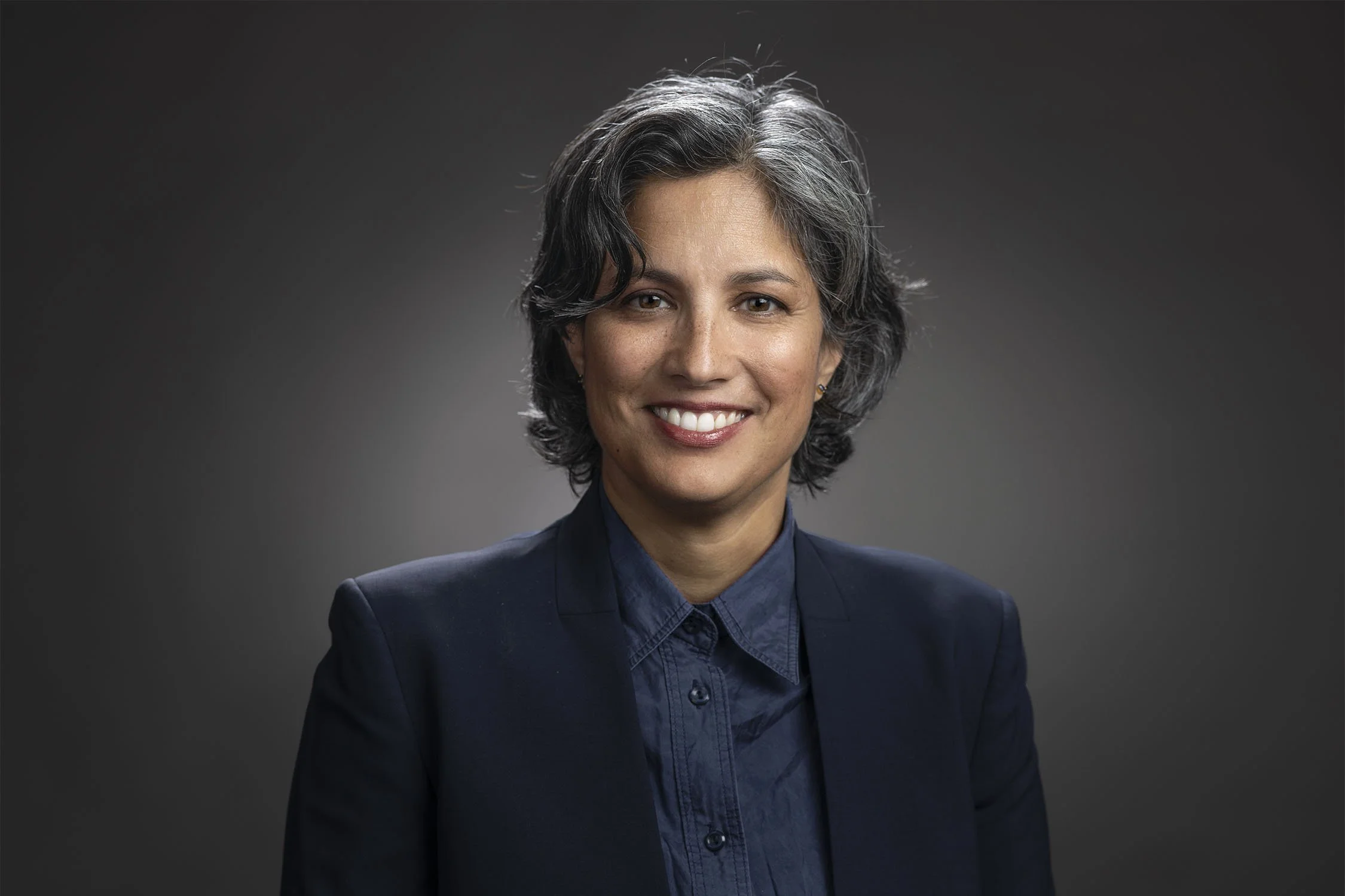 Professional portrait of a smiling woman with short gray hair, wearing a dark suit jacket and a navy button-up shirt, against a dark gray background.