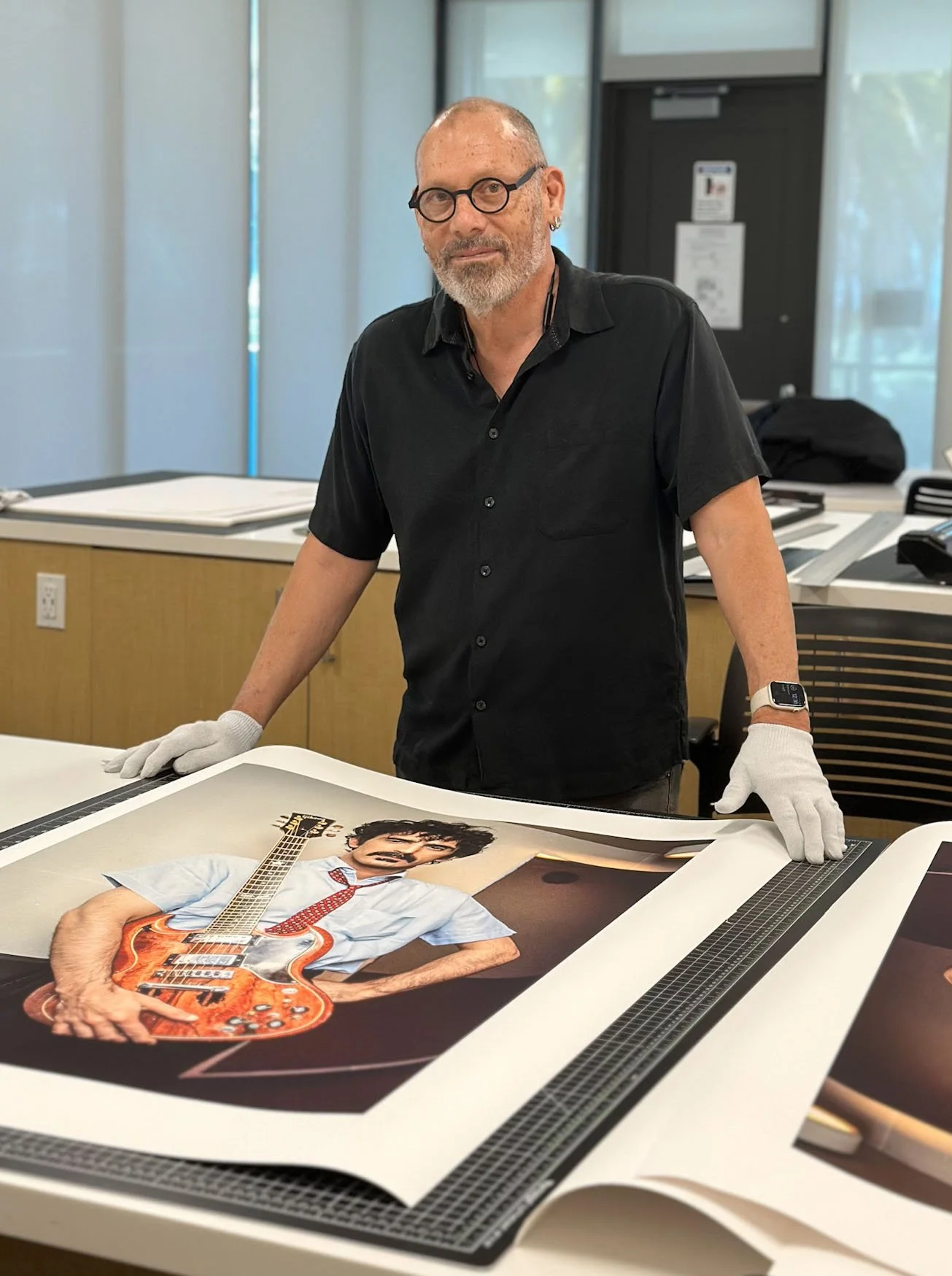 A man wearing glasses, a black shirt, and white gloves standing in an office, displaying rolled large color prints of a person holding an electric guitar.