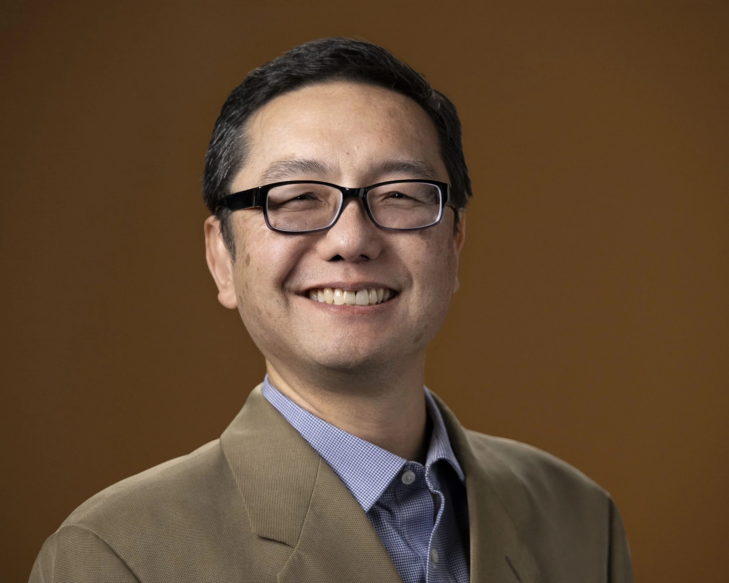 A smiling man with black hair, glasses, wearing a beige suit jacket and a light blue collared shirt, against a brown background.