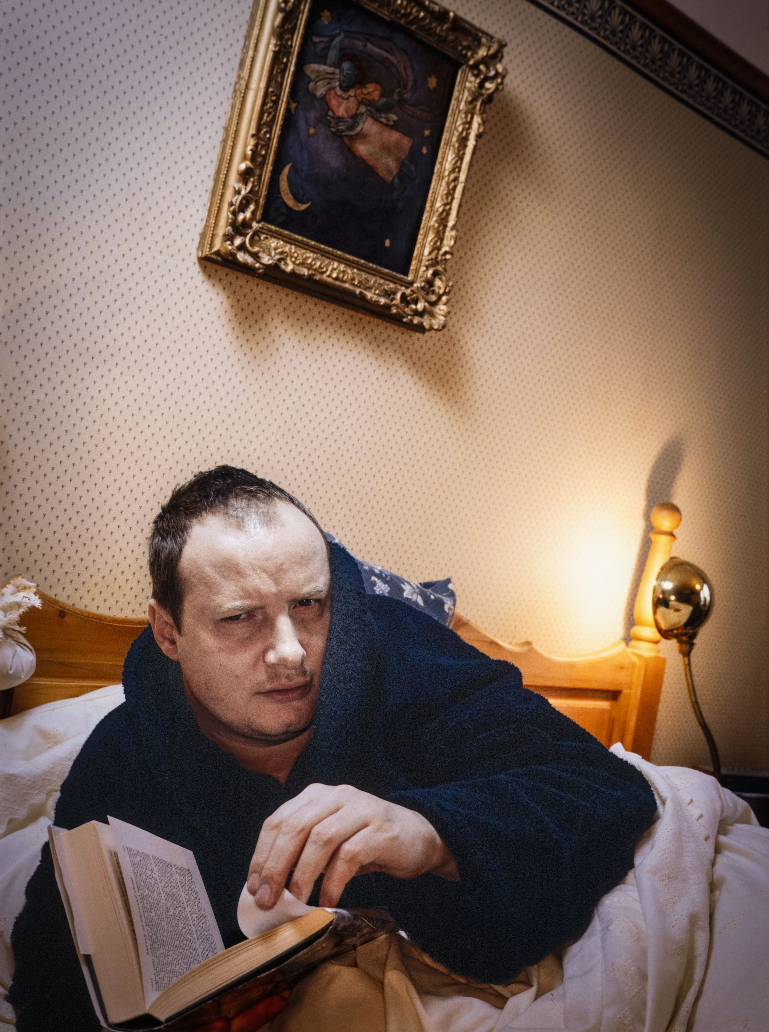 A man lying in bed, wearing a dark blue robe,, reading a book with a puzzled or suspicious expression. The room has beige dotted wallpaper, a wooden headboard, a lamp, and framed artwork of a woman flying through the sky with stars and a crescent moon.