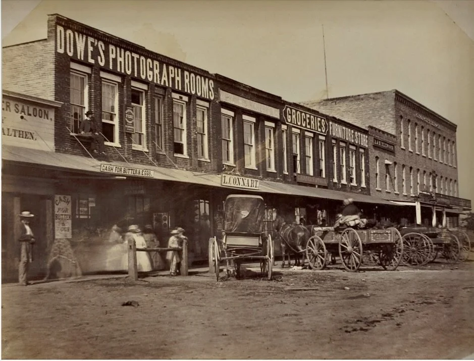 An old image of horse drawn wagons in front of brick and mortar stores.