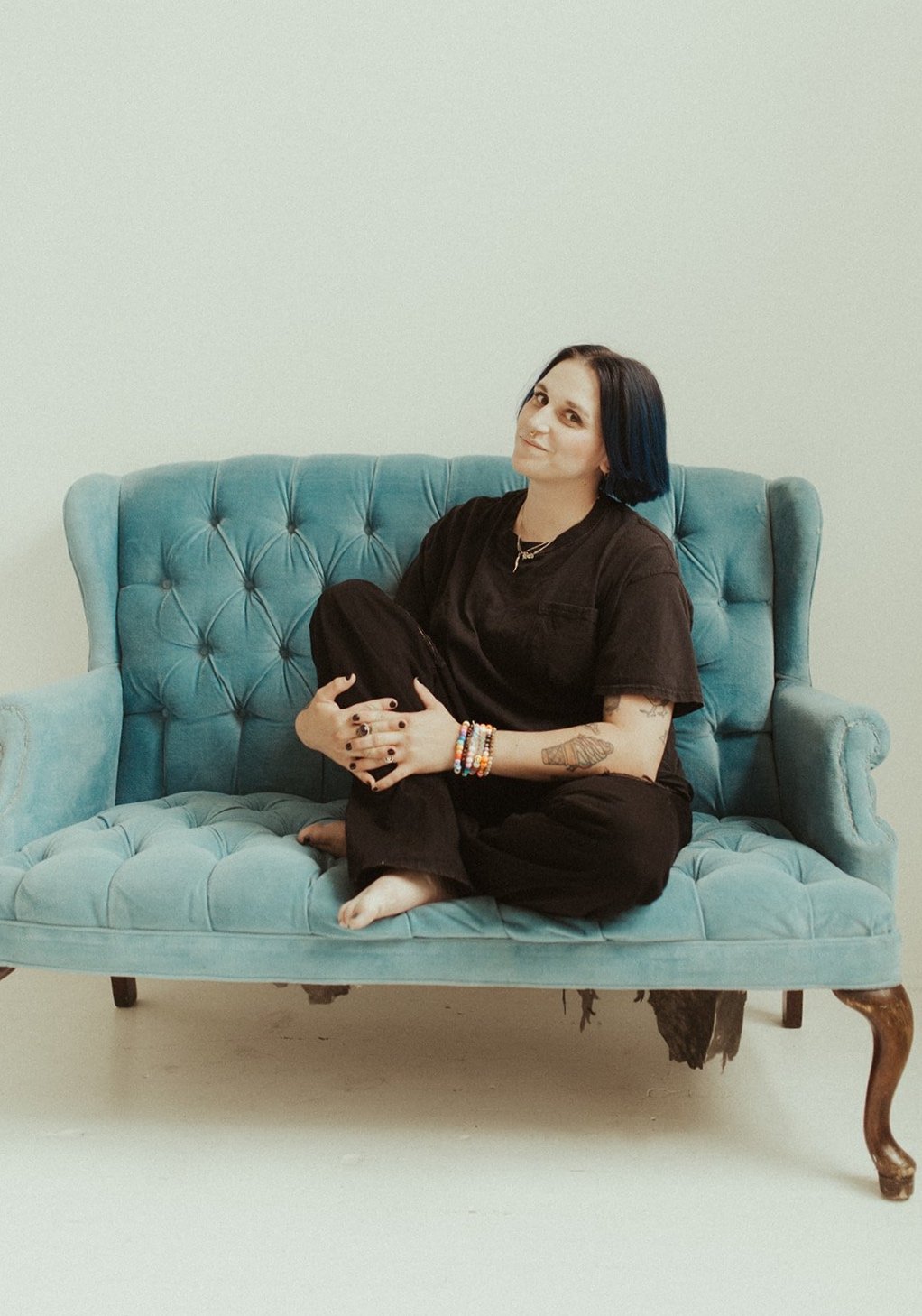 A woman with black hair and tattoos, dressed in black, sitting barefoot on a vintage teal sofa, smiling and holding a black purse against a plain white wall.