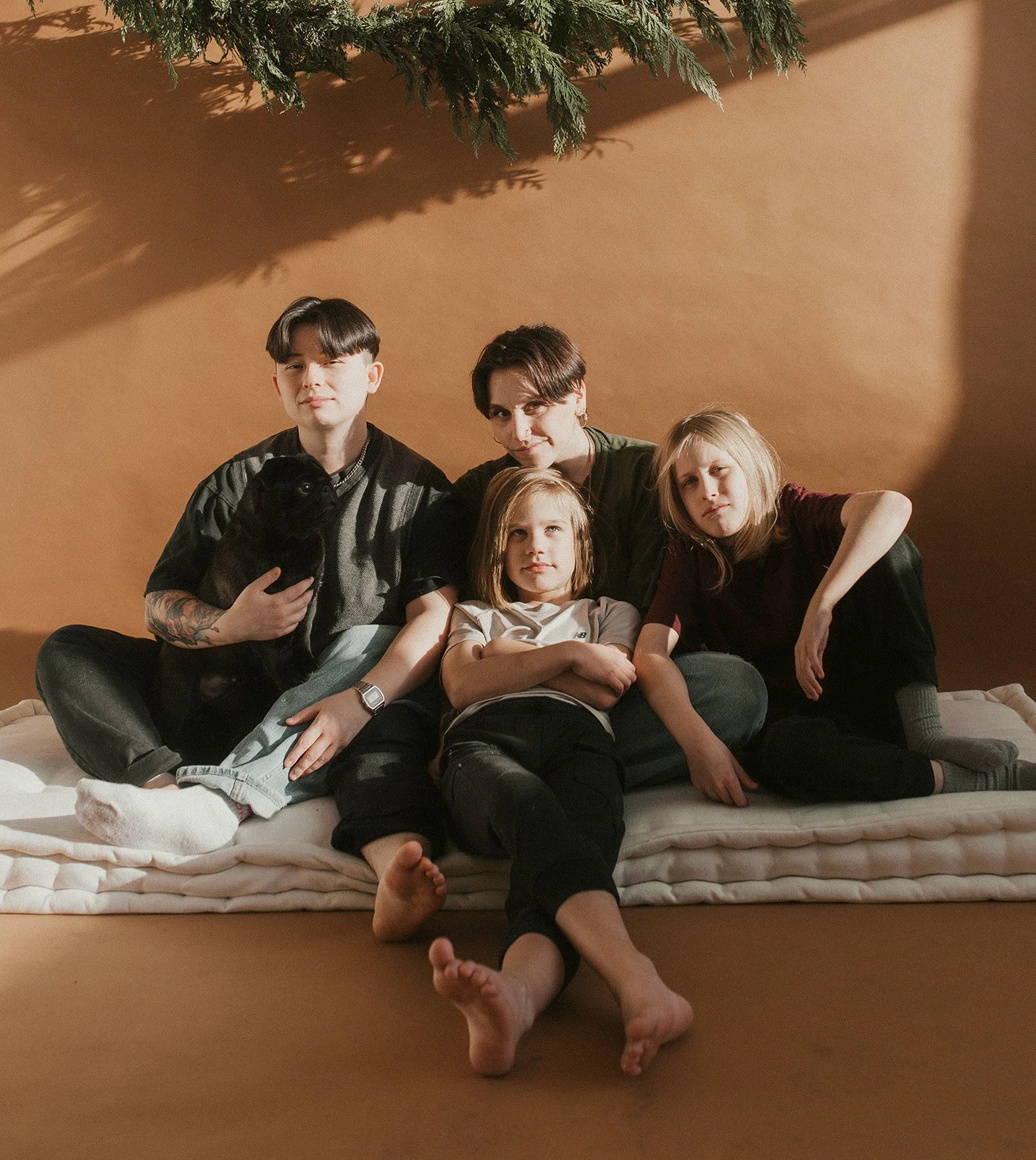 Four young people, including children and adults, sitting on a mattress with relaxed and playful expressions, a black puppy on one person's lap, and a Christmas tree branch visible at the top, against a warm-toned background.