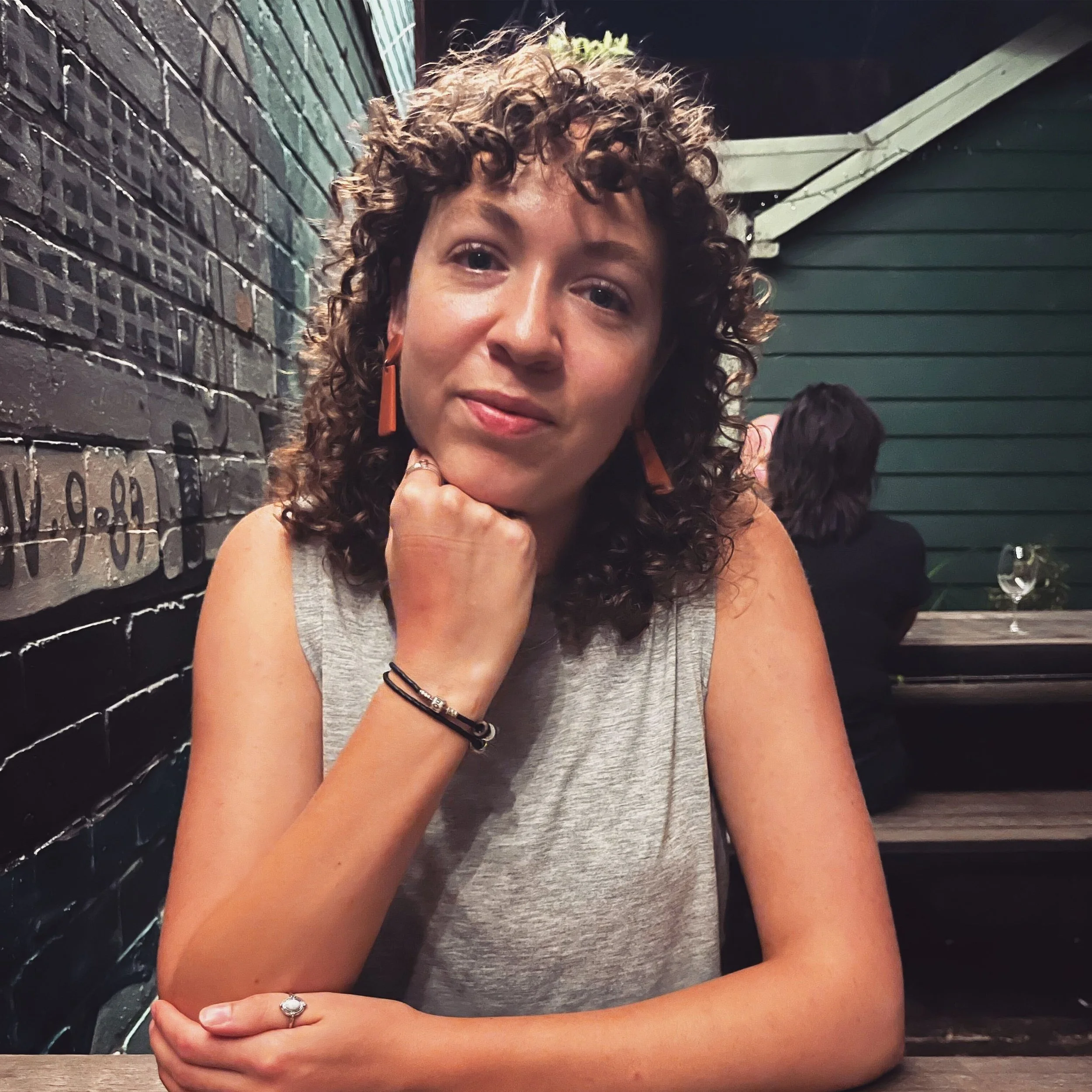 Photograph of Emily Pearce, she's sat at a wooden table. She's got brown curly hair, orange earrings and a grey vest top on. She's resting her chin on her knuckles.