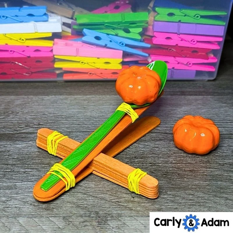 Close-up of a pumpkin catapult made from orange craft sticks, yellow rubber bands, and a green plastic spoon. One orange pumpkin rests in the spoon and another sits on the gray tabletop.