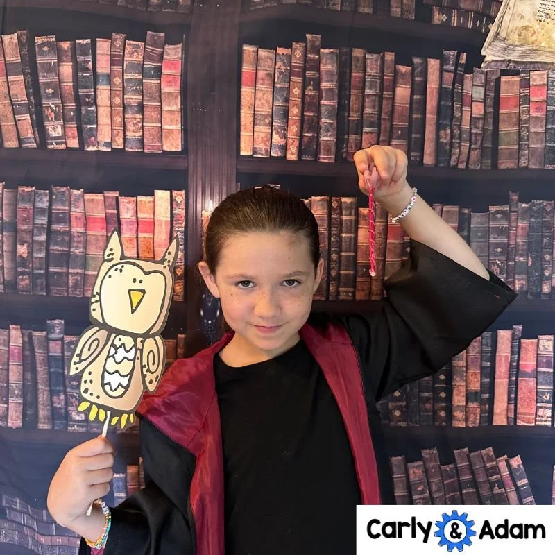 A child wearing a black robe with red lining holds an owl photo prop and raises a wand in front of a backdrop printed with old books.