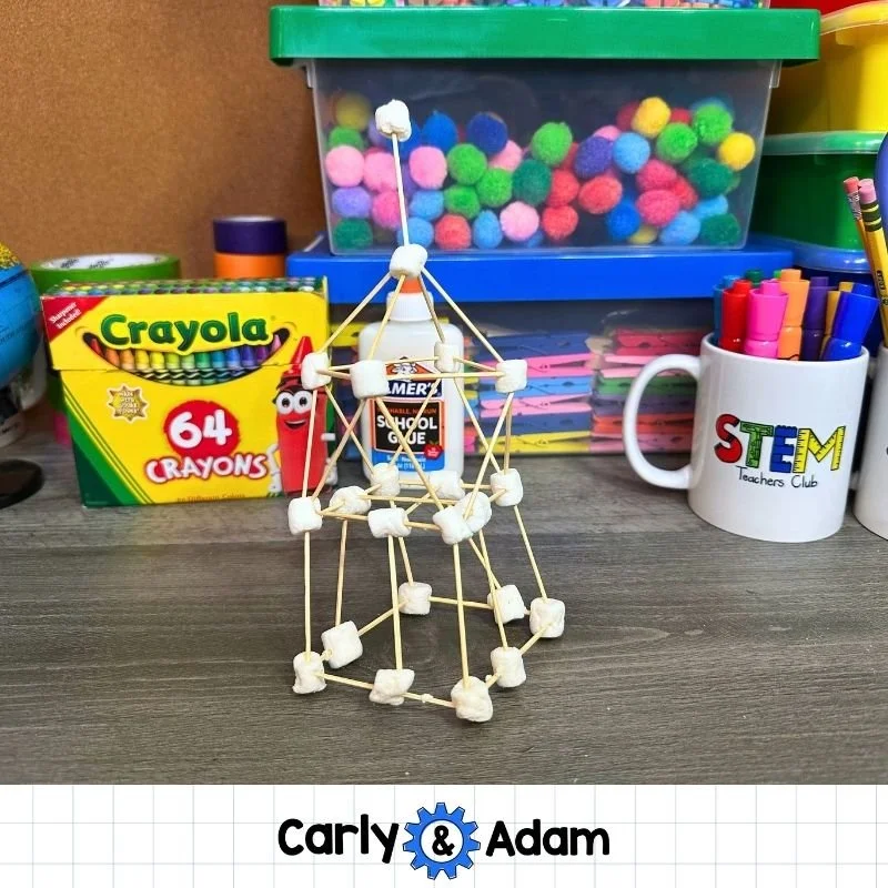 A tall toothpick tower connected with white marshmallows stands on a classroom table. The structure has several levels, diagonal supports, and a pointed top.
