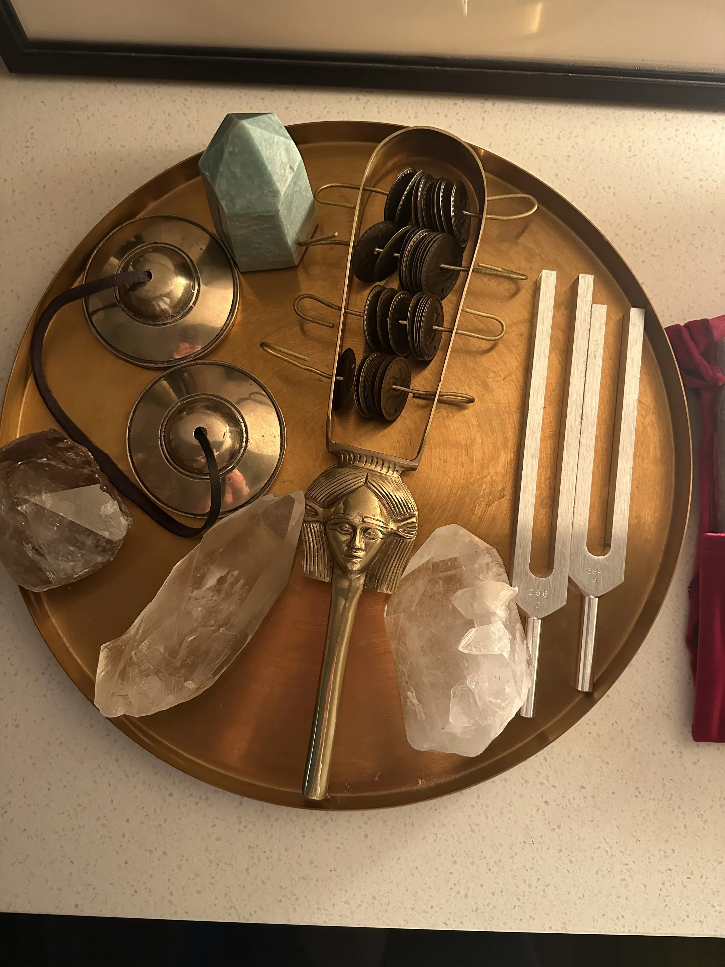 A round golden tray holds a brass ceremonial Egyptian sistrum, tingsha bells, tuning forks, and several large clear quartz crystals, with a partial view of a framed Jikiden Reiki Institute Shoden and Okuden certification in Japanese behind.