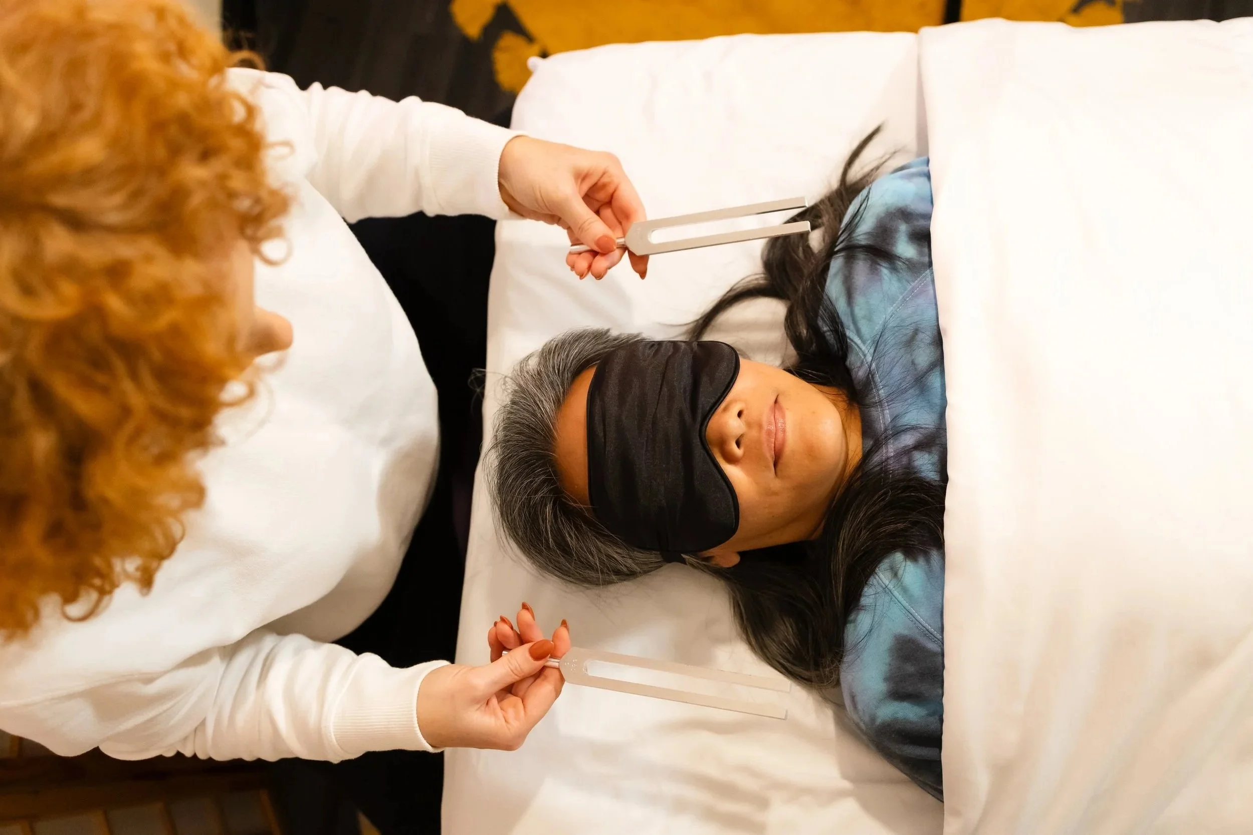 A woman wearing a sleep mask lies on a white bed, while another woman with red curly hair holds tuning forks at both sides of her head