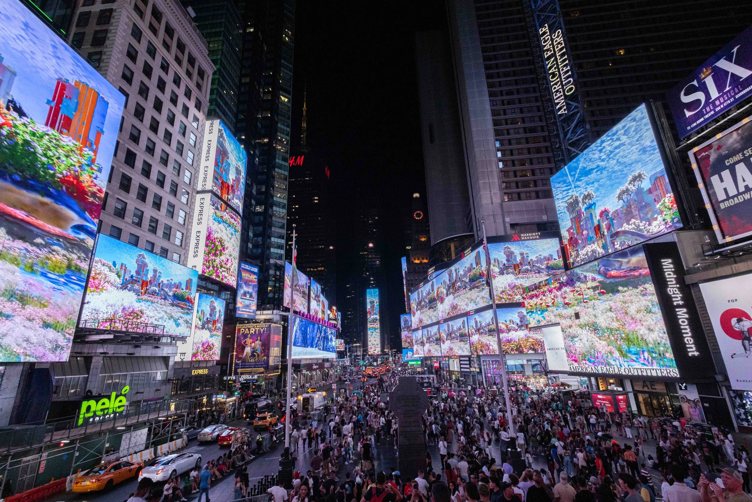 A crowded city street at night with bright digital billboards and advertisements, including a colorful display of flowers and a marquee for the musical 'Aladdin'.