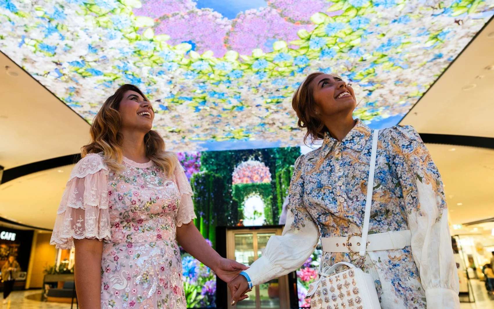 Two women joyfully holding hands and smiling inside a shopping mall, with a vibrant floral canopy overhead and a digital screen showing a garden scene behind them.