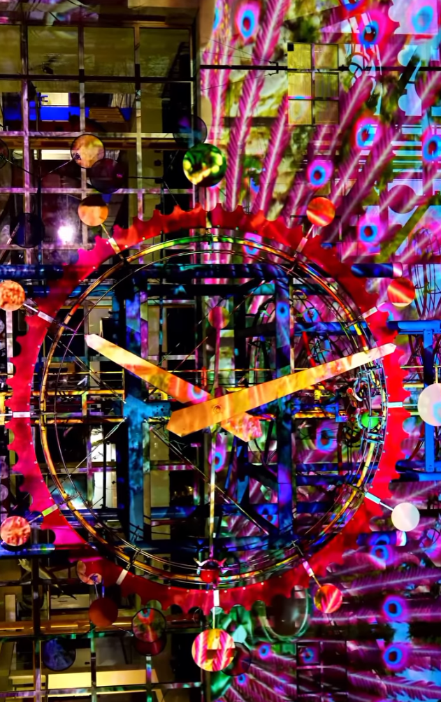 Colorful abstract art installation with a large clock face at the center, surrounded by vibrant patterns, geometric shapes, and multicolored spheres.