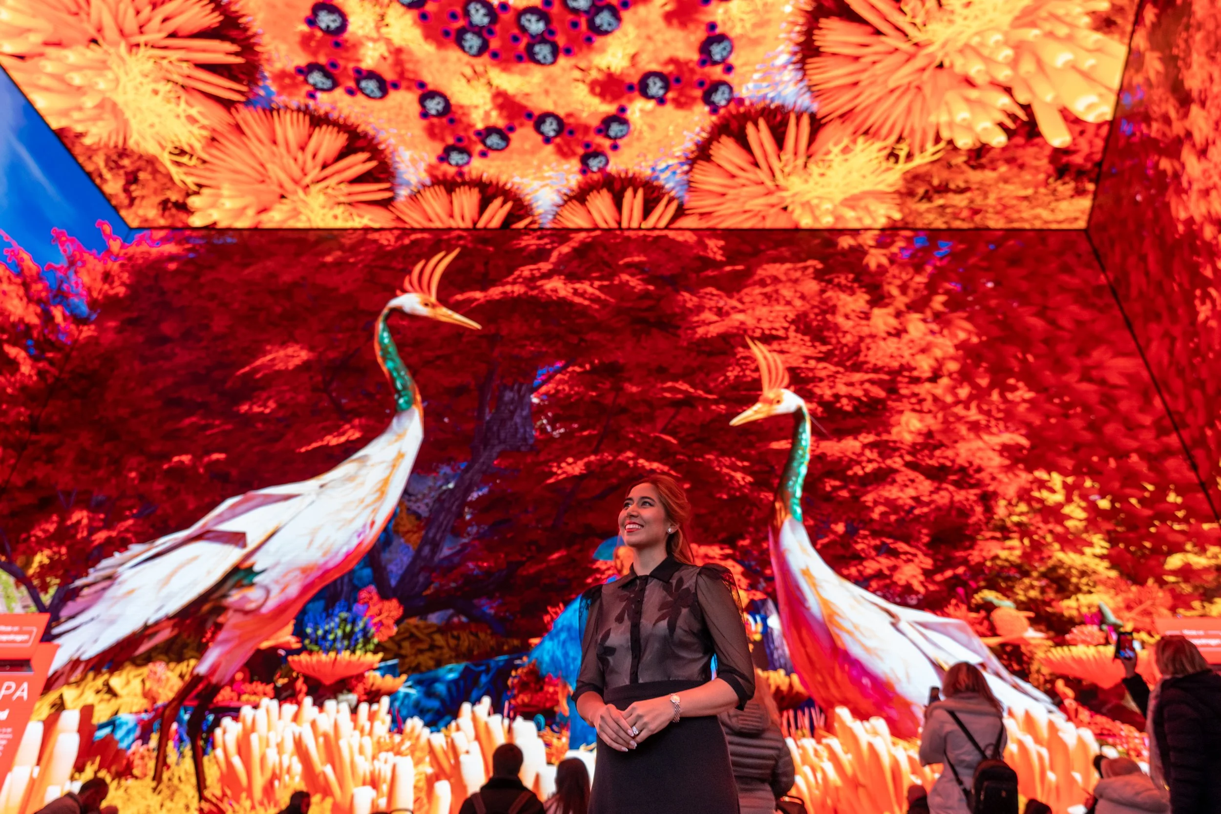 A woman standing amidst an elaborate indoor digital art display featuring large, colorful representations of peacocks, trees, and flowers, with people in the background enjoying the exhibit.