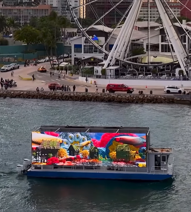 A floating digital billboard displays colorful artwork with dragons and flowers on the water near a waterfront walkway, in front of an amusement park with a large Ferris wheel and buildings.