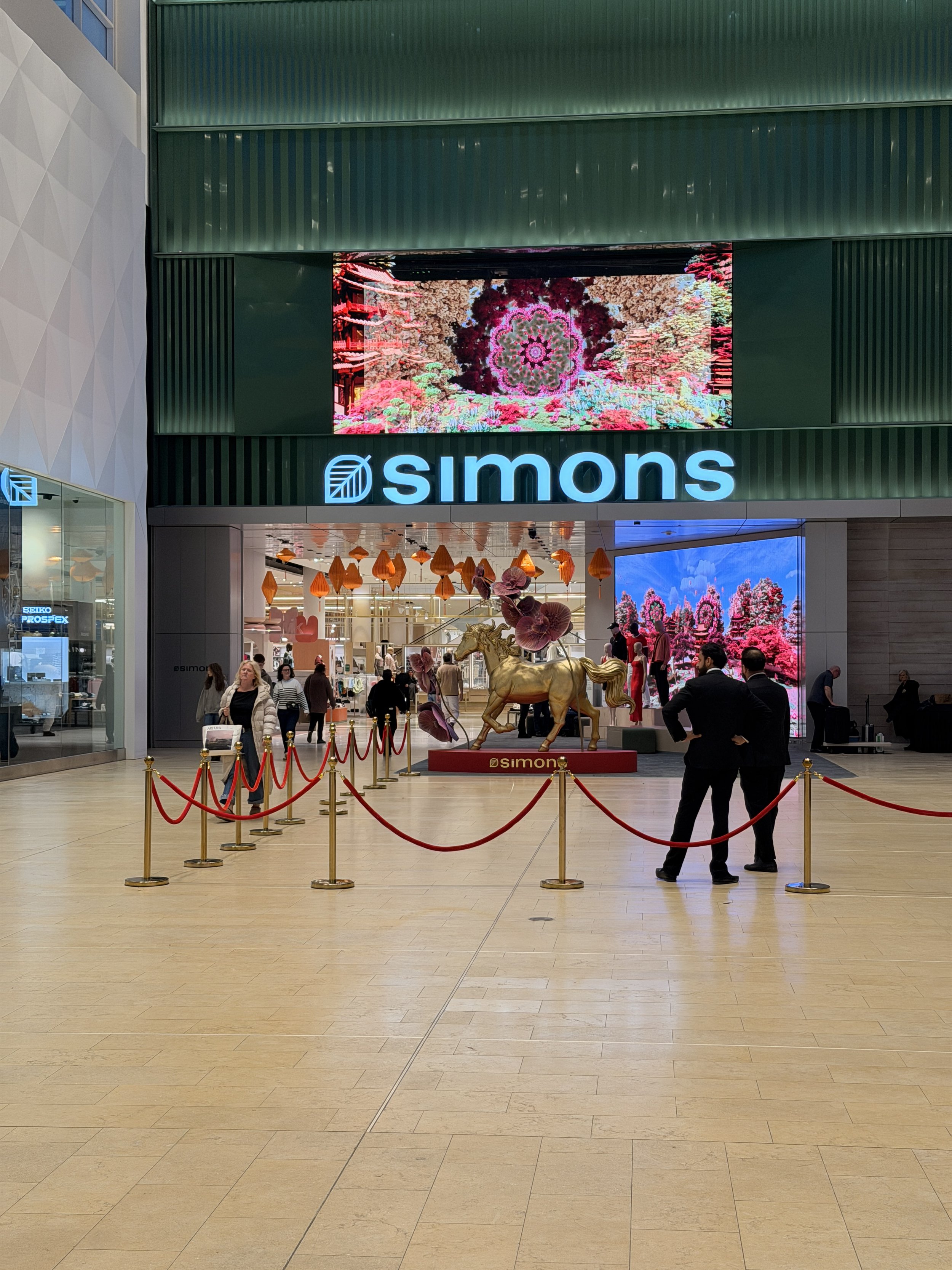 Inside a shopping mall with people walking and shopping. There is a large digital screen displaying a colorful floral pattern above the storefront of Simons, a department store. In the foreground, a golden unicorn statue with pink flowers around its neck is up on a red platform. Red ropes are set up to create a queuing area around the statue. Several shoppers are near the entrance, some looking at the display and some talking.