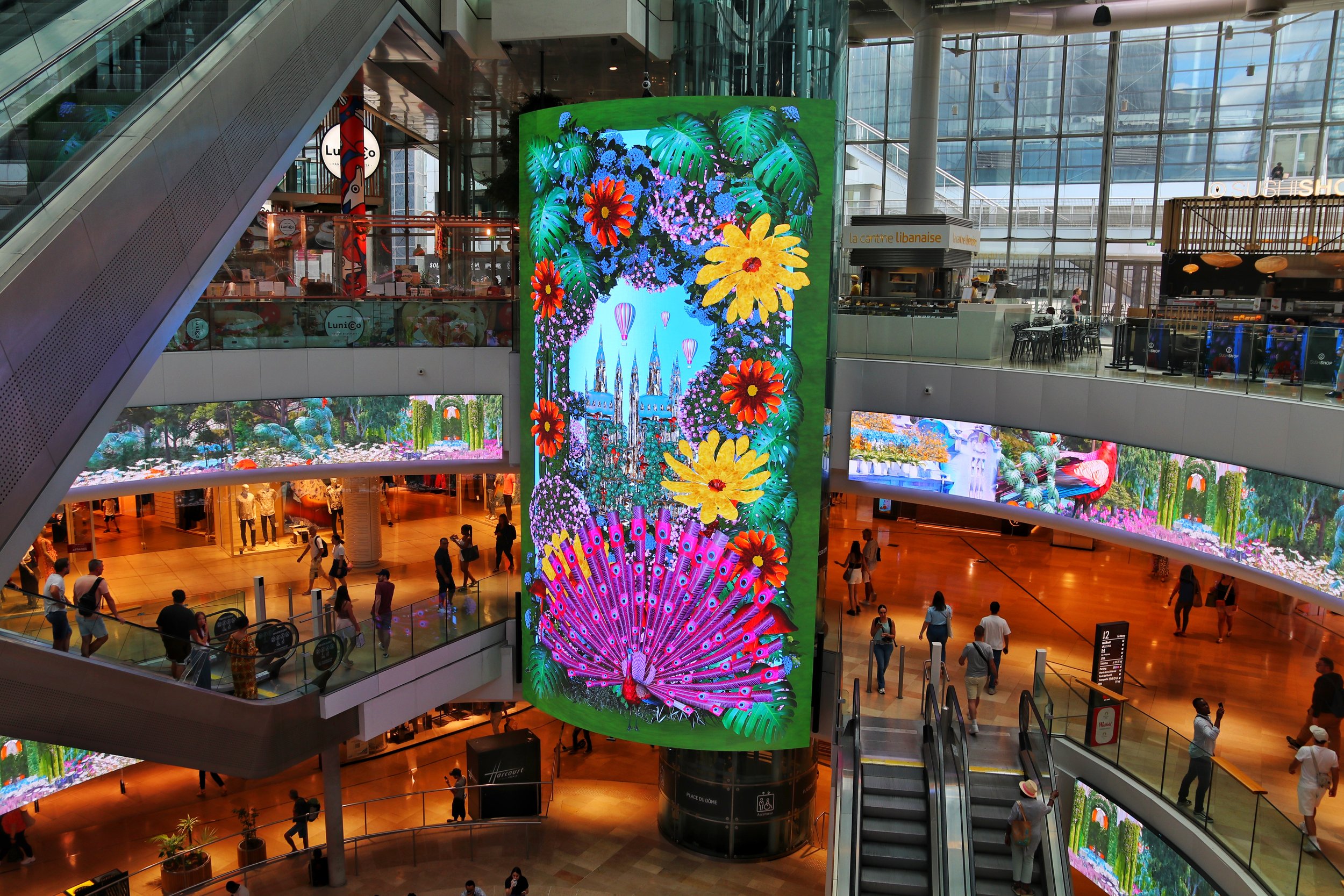 Interior of a modern shopping mall with multiple levels, escalators, and large digital display screens showcasing colorful floral and scenic images. People are walking around and shopping.