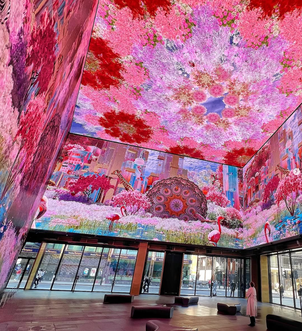 The interior of a modern building with large glass windows, featuring a digital art display of a vibrant, colorful fantasy cityscape with pink trees, flamingos, skyscrapers, a giraffe, and a decorative mandala on the ceiling.