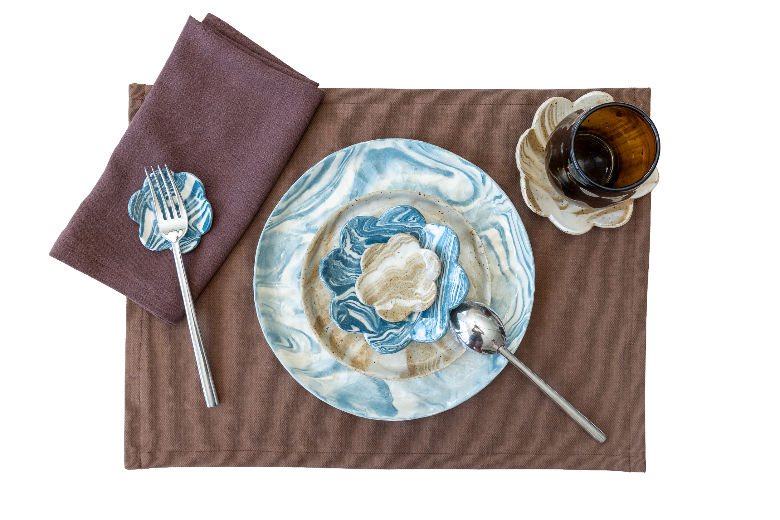 Marbled blue, white, and beige ceramic plate with a decorative flower-shaped dish holding cookies or crackers, a brown napkin with a fork on top, a spoon, and a brown glass cup on a saucer, all arranged on a brown placemat.