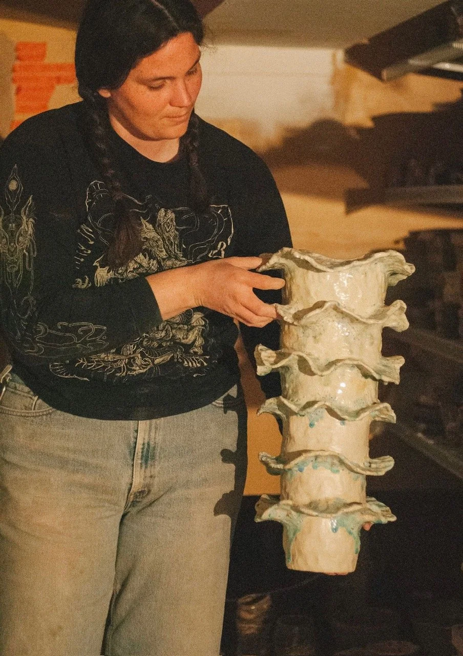 A woman with dark hair styled in two braids holding a large ceramic sculpture of a human spine with visible vertebrae.