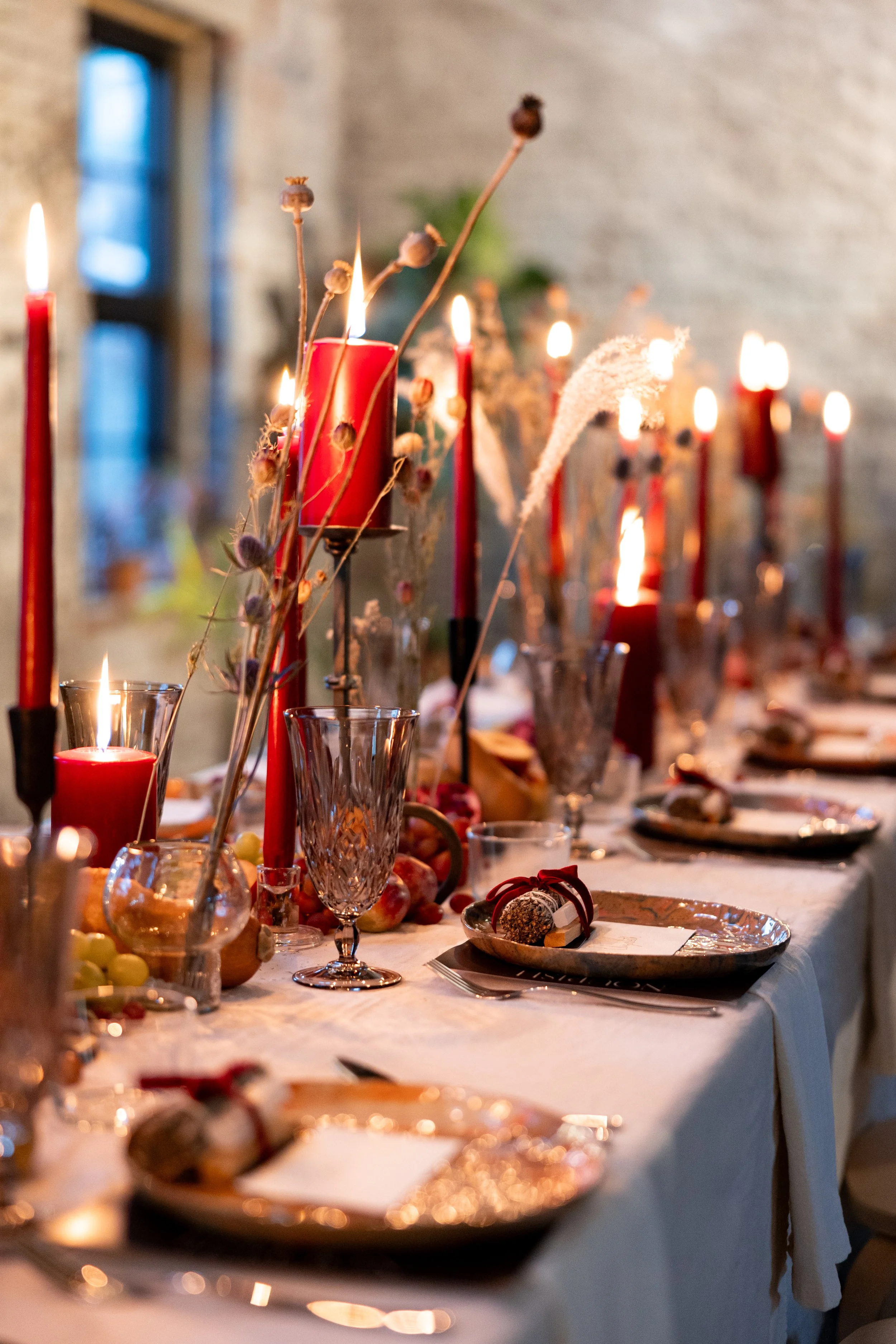 A festive dinner table with lit red candles, glassware, plates, and decorative elements. The background features a brick wall and a window with natural light.