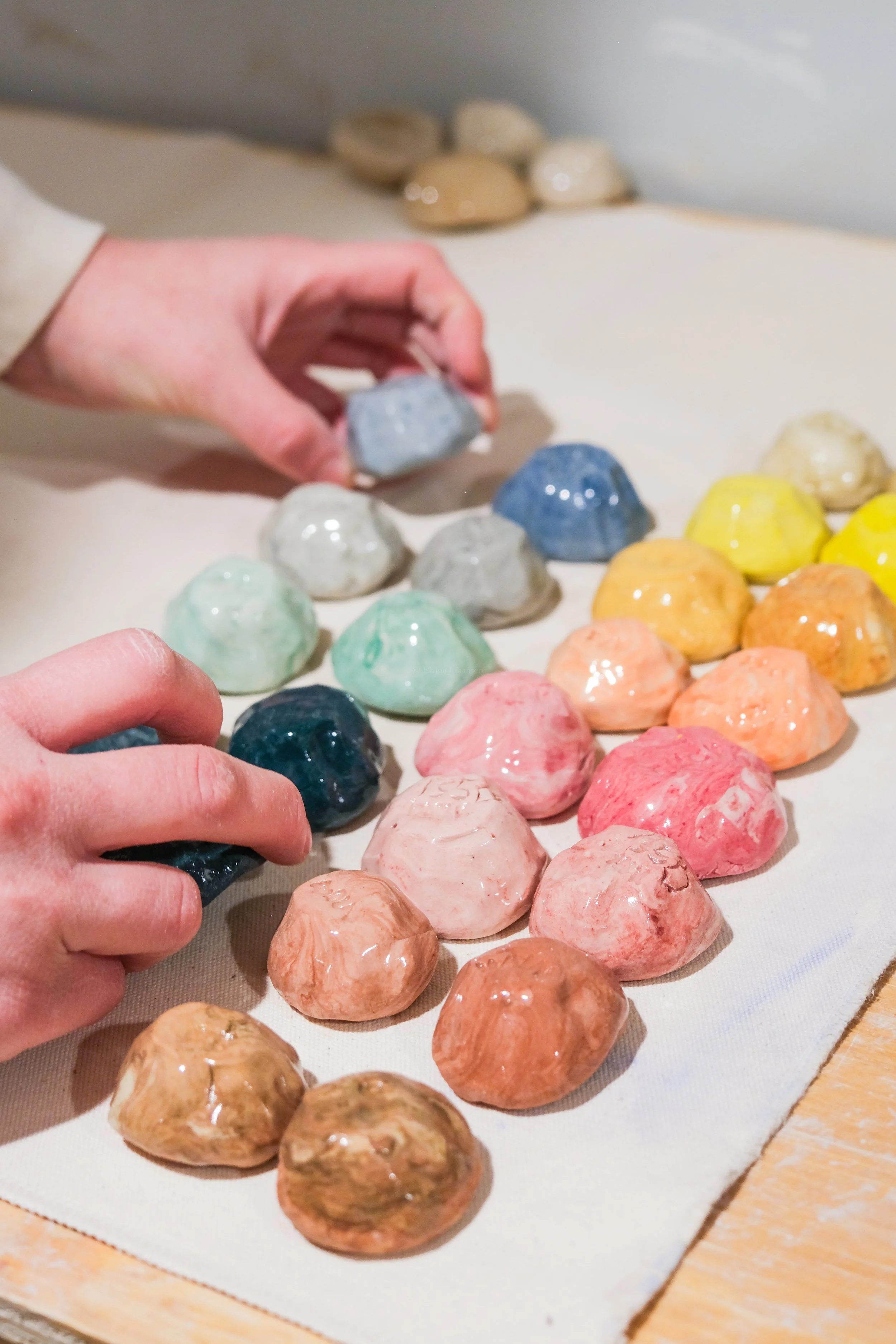 A person is placing colorful ceramic shells on a white cloth. The shells are arranged in rows and come in various pastel and bright colors, including blue, green, orange, pink, and yellow.