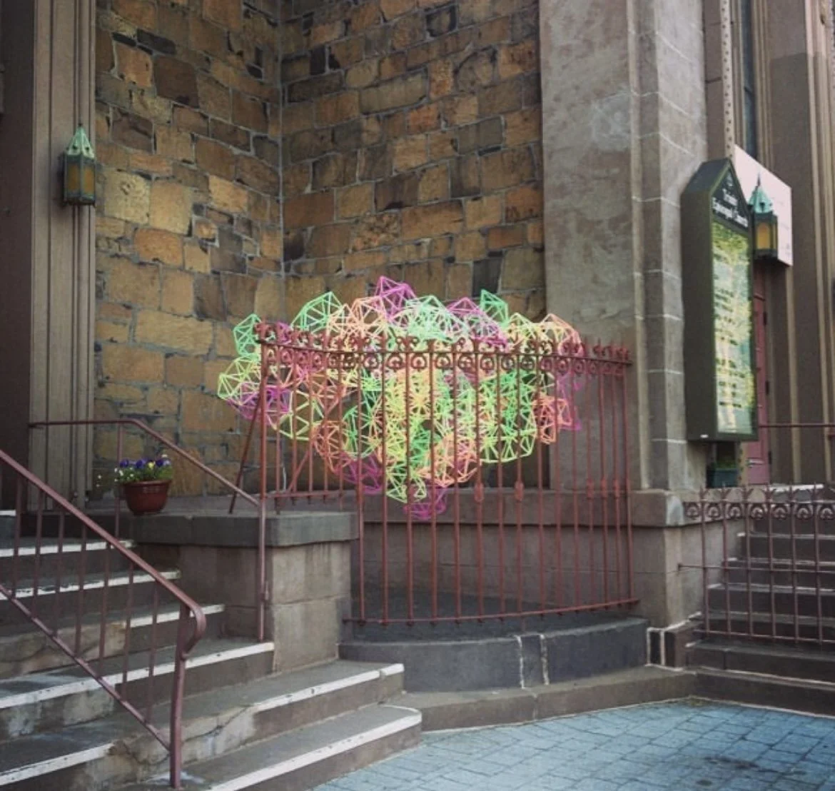 Colorful geometric light installation resembling a flower or starburst, set against a brick building exterior with stairs and a metal railing.