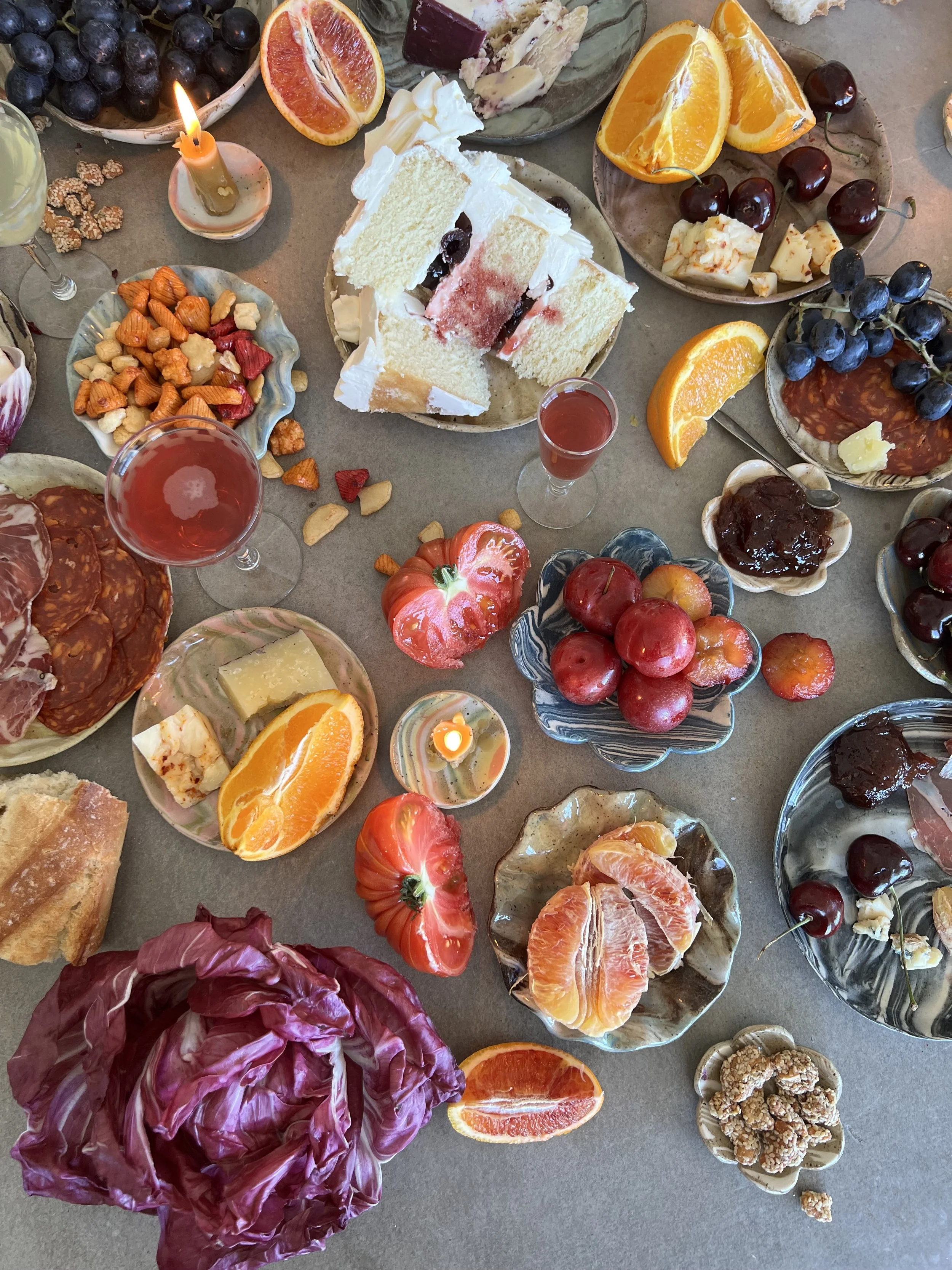 An assortment of food on a table, including sliced oranges, grapes, cherries, a slice of watermelon, a cake with white frosting, small bowls of cheese, nuts, and jam, a lit candle, and various other snacks and fruits.