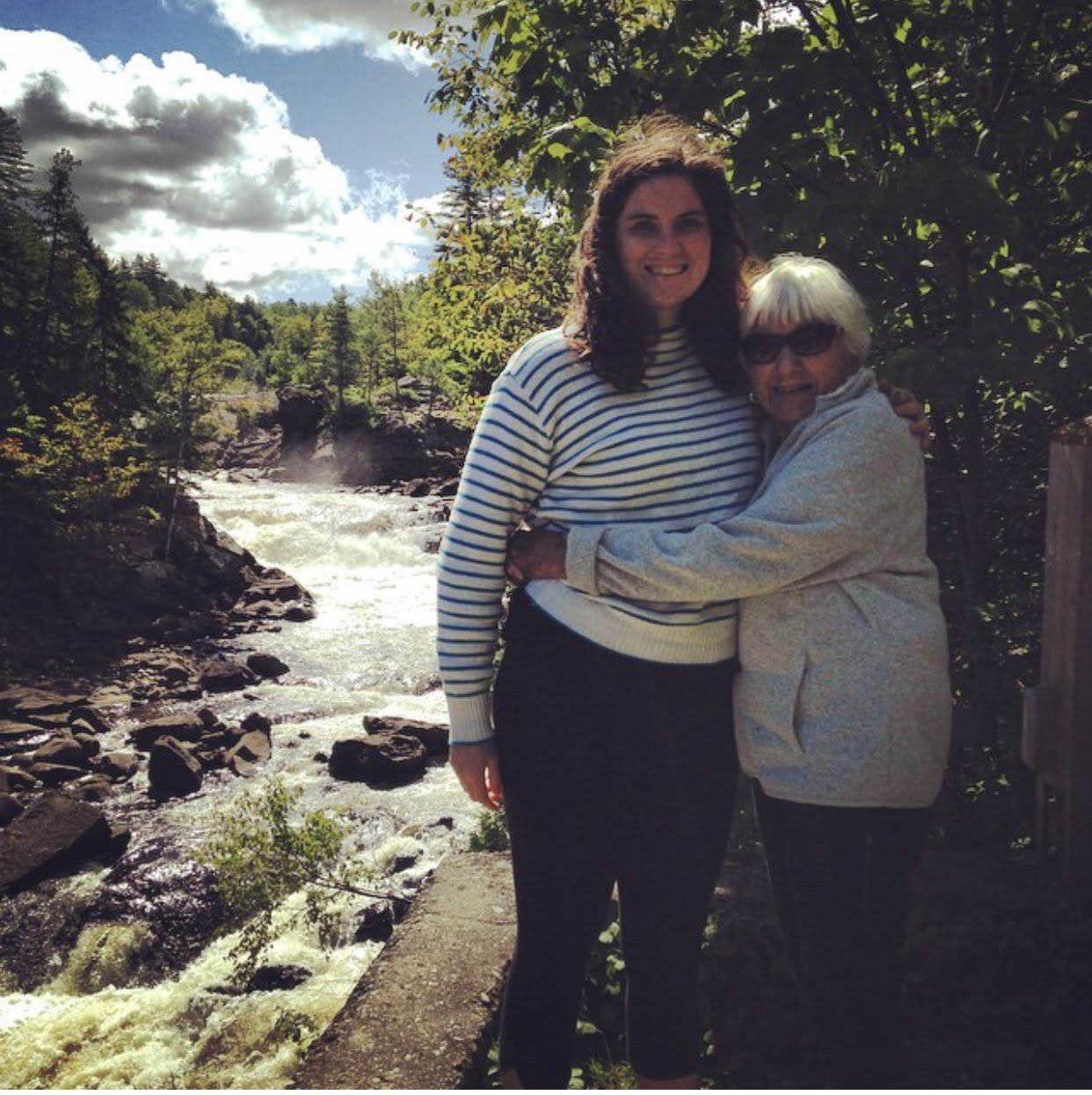 Two women embracing outdoors near a river with trees and cloudy sky in the background.