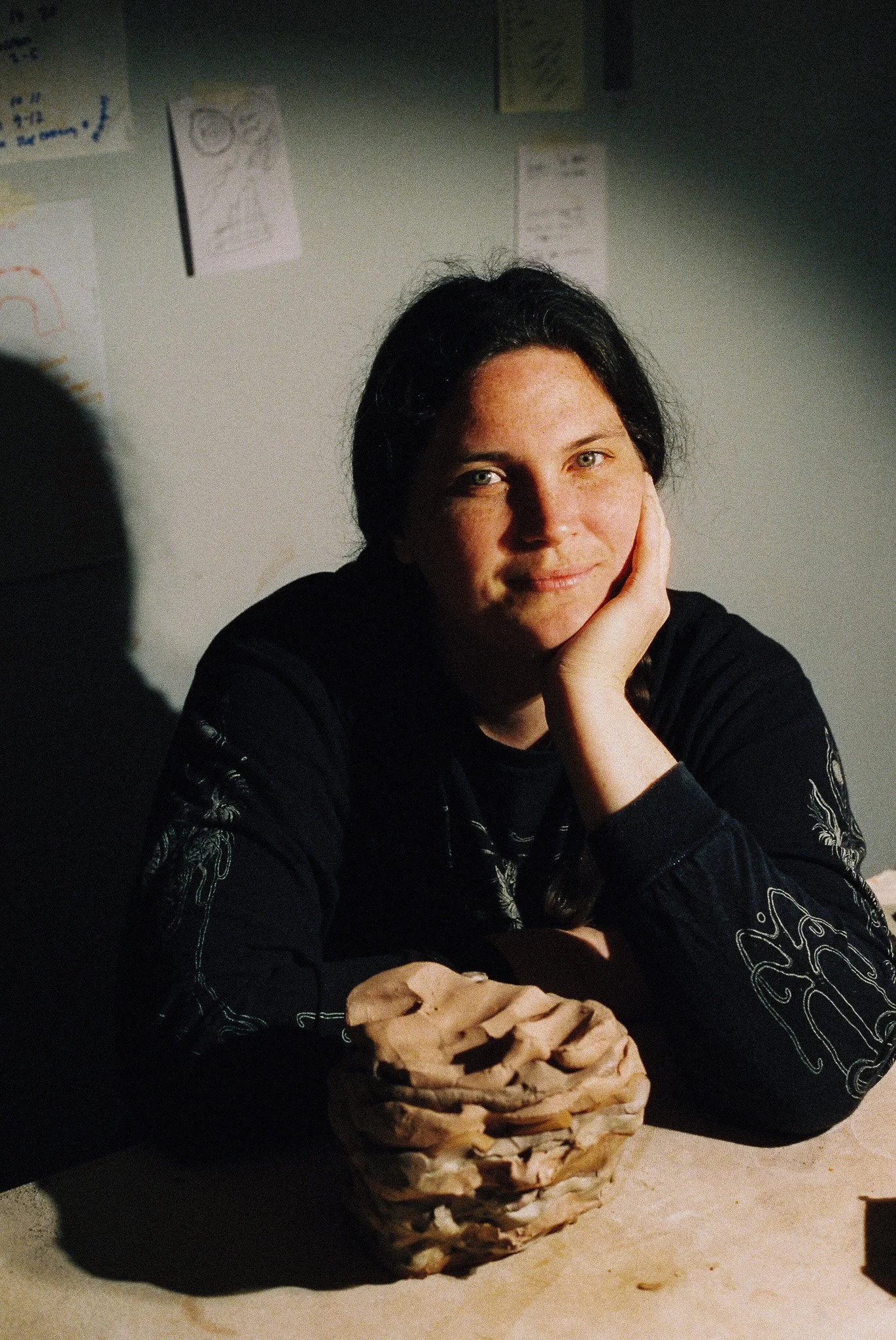 A woman with black hair and light skin, resting her head on her hand, sitting at a table with a large, carved wooden sculpture in front of her. The background features a wall with various sketches and notes.