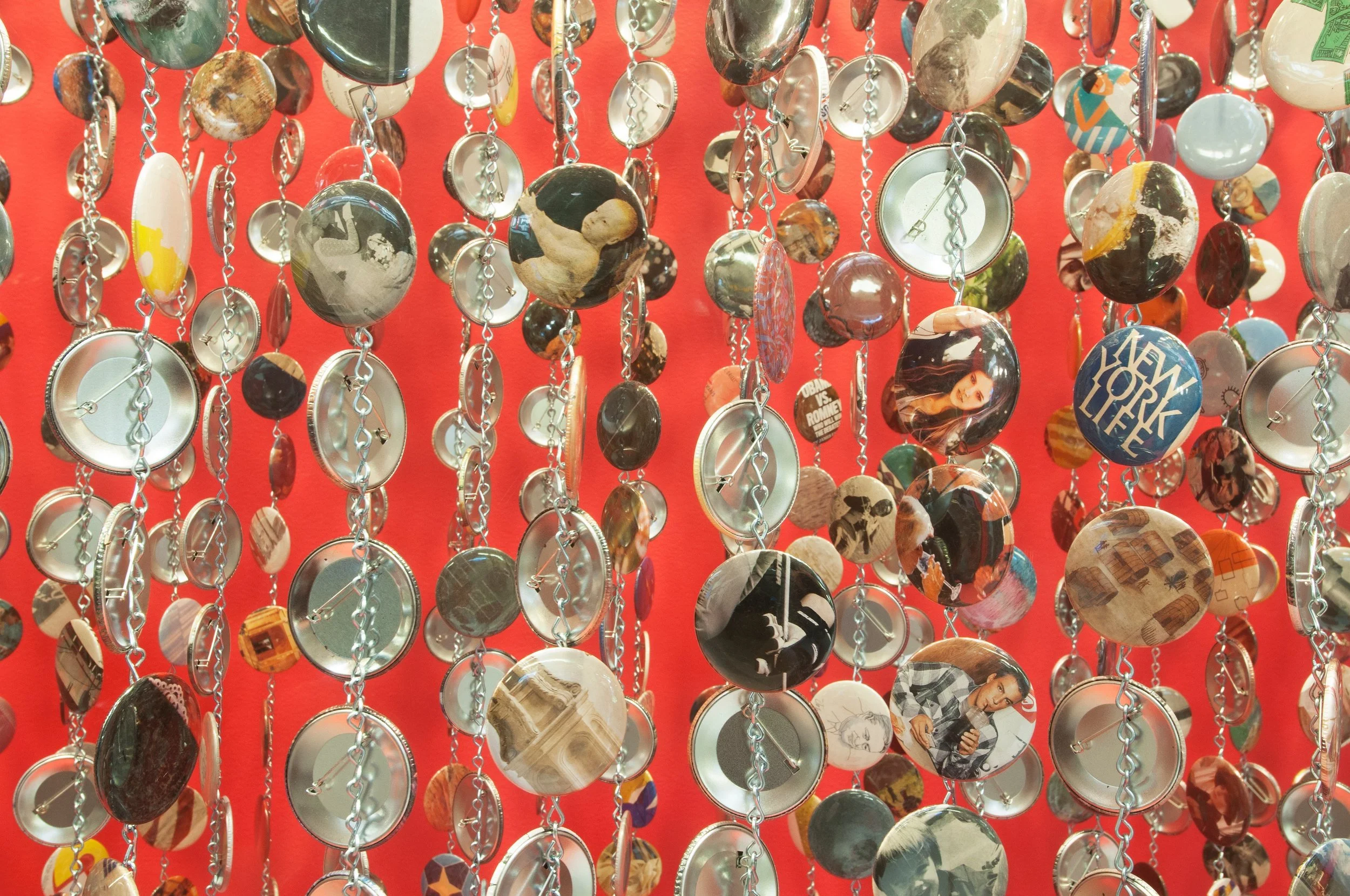 A hanging art installation made of numerous circular elements attached by chains. The circles feature various images, including people, artwork, and text, set against a bright red background.