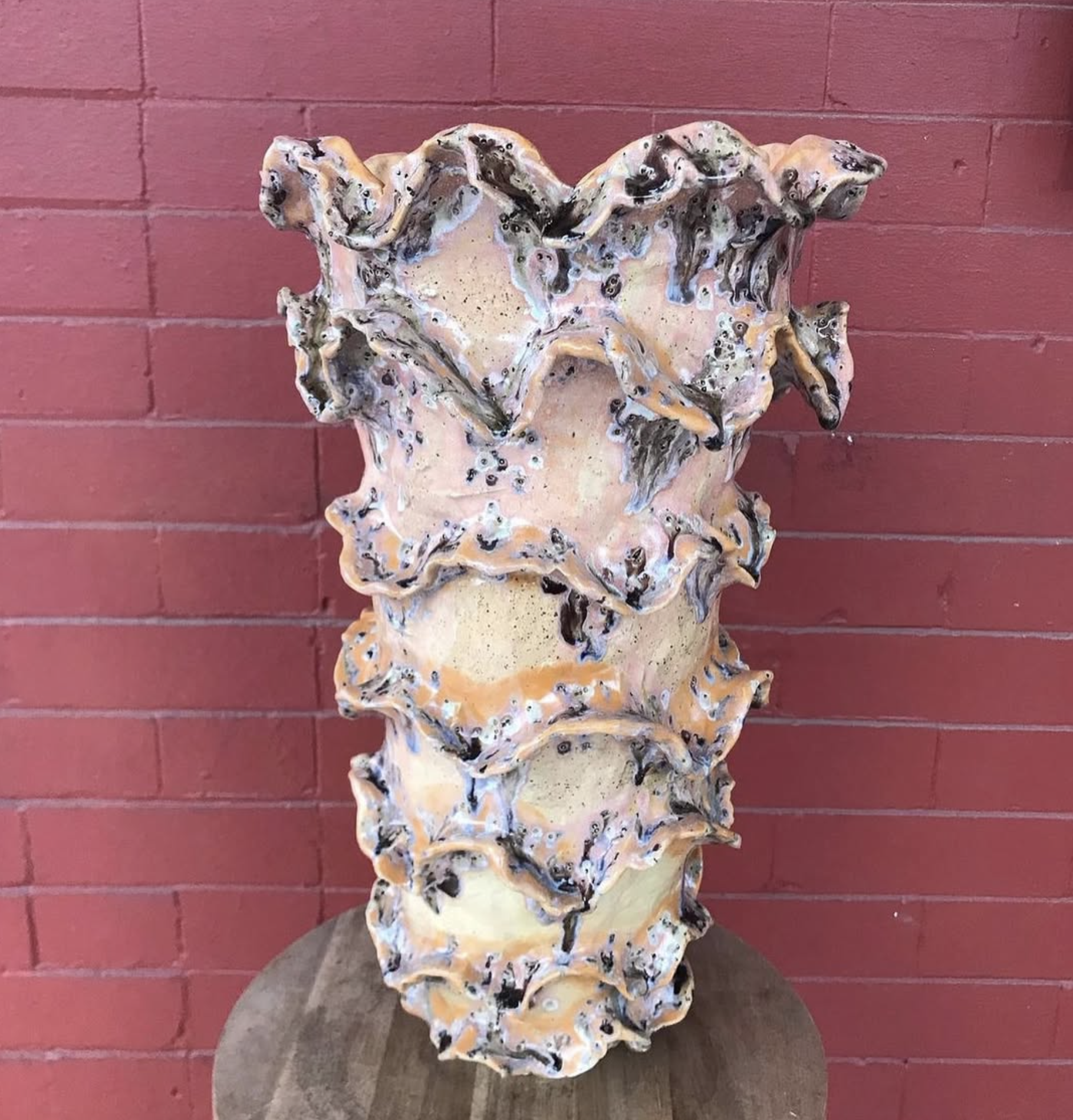A decorative ceramic or plaster sculpture of a human face with a textured and wavy design, mounted on a stone or concrete pedestal against a red brick wall.
