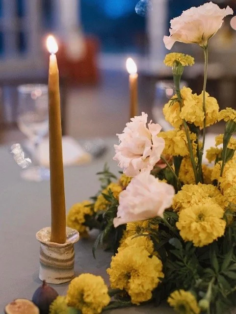 A table centerpiece with yellow and pink flowers and lit candles at a dinner setting.