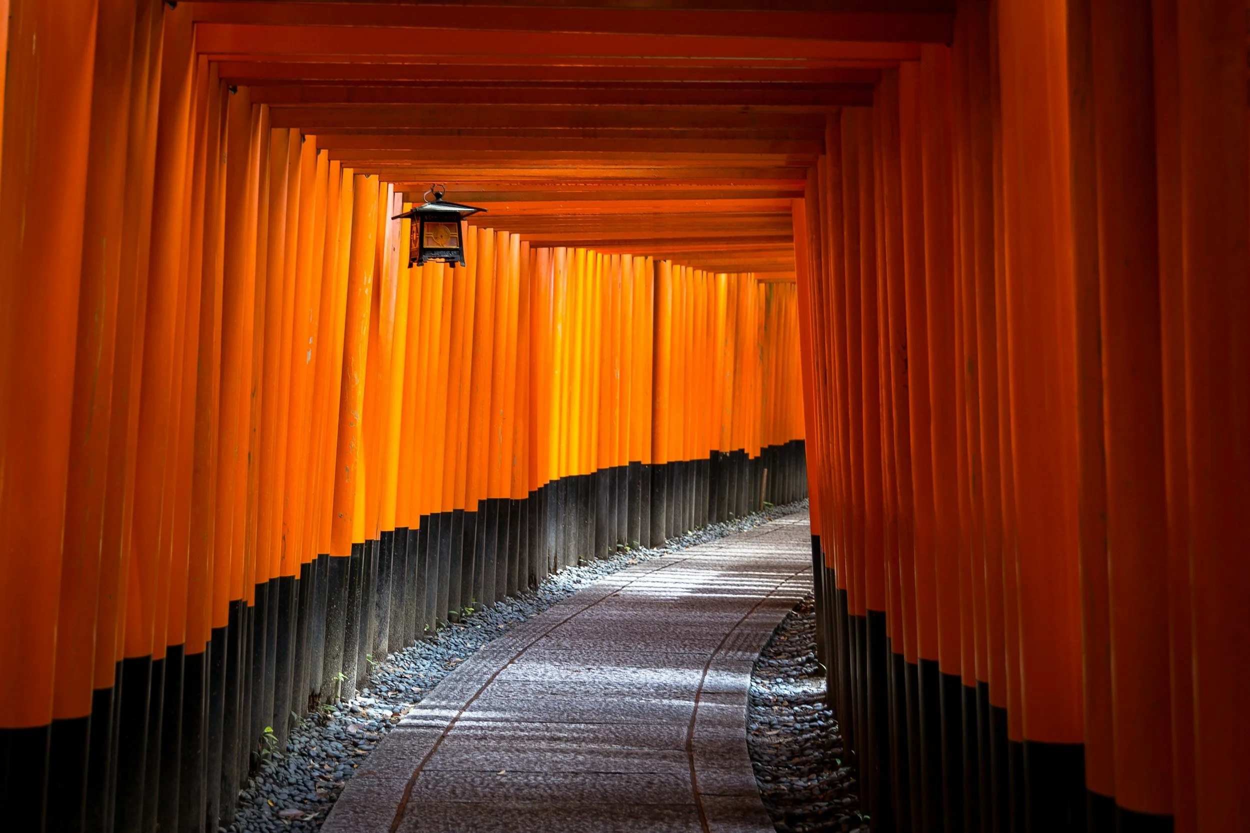 A pathway through a tunnel of orange torii gates at Fushimi Inari Shrine, with a small lantern hanging from the ceiling.