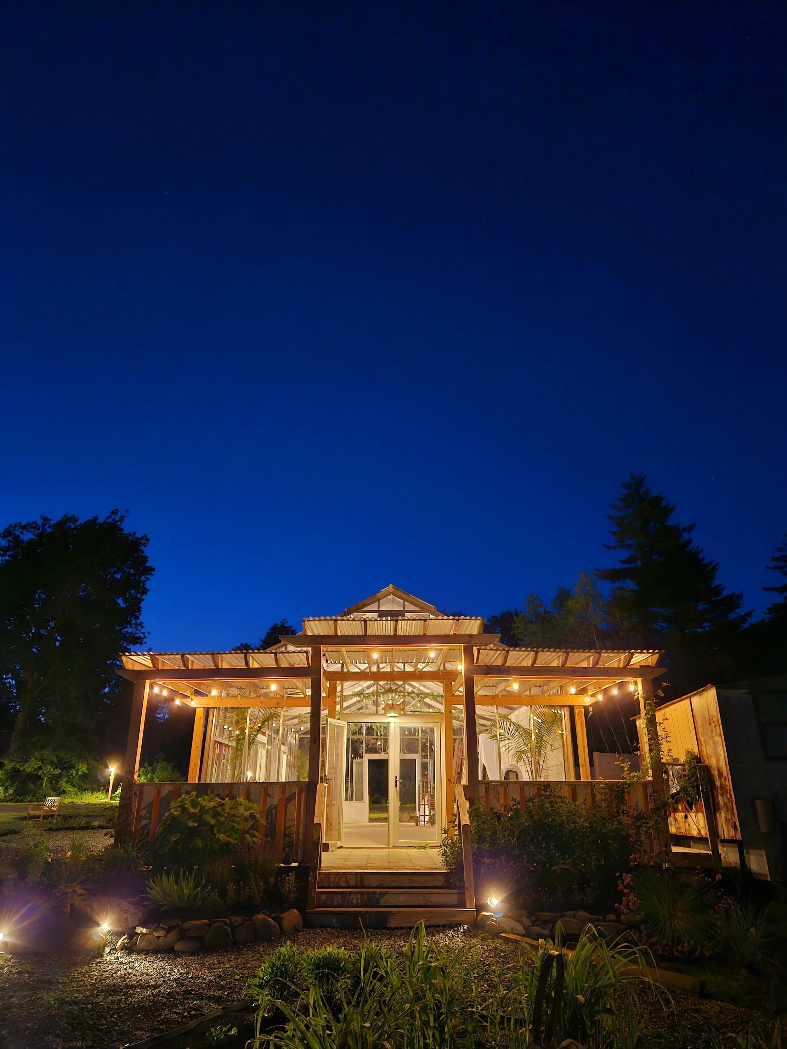 A warmly lit wooden house with a porch at night, surrounded by trees and garden, under a clear dark blue sky.