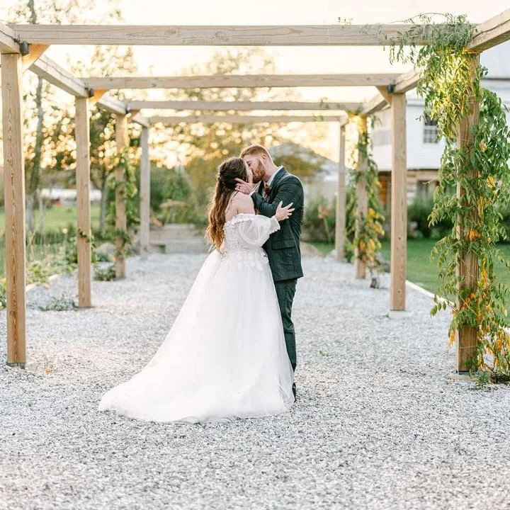 A bride and groom sharing a kiss under a wooden pergola during their outdoor wedding at sunset.