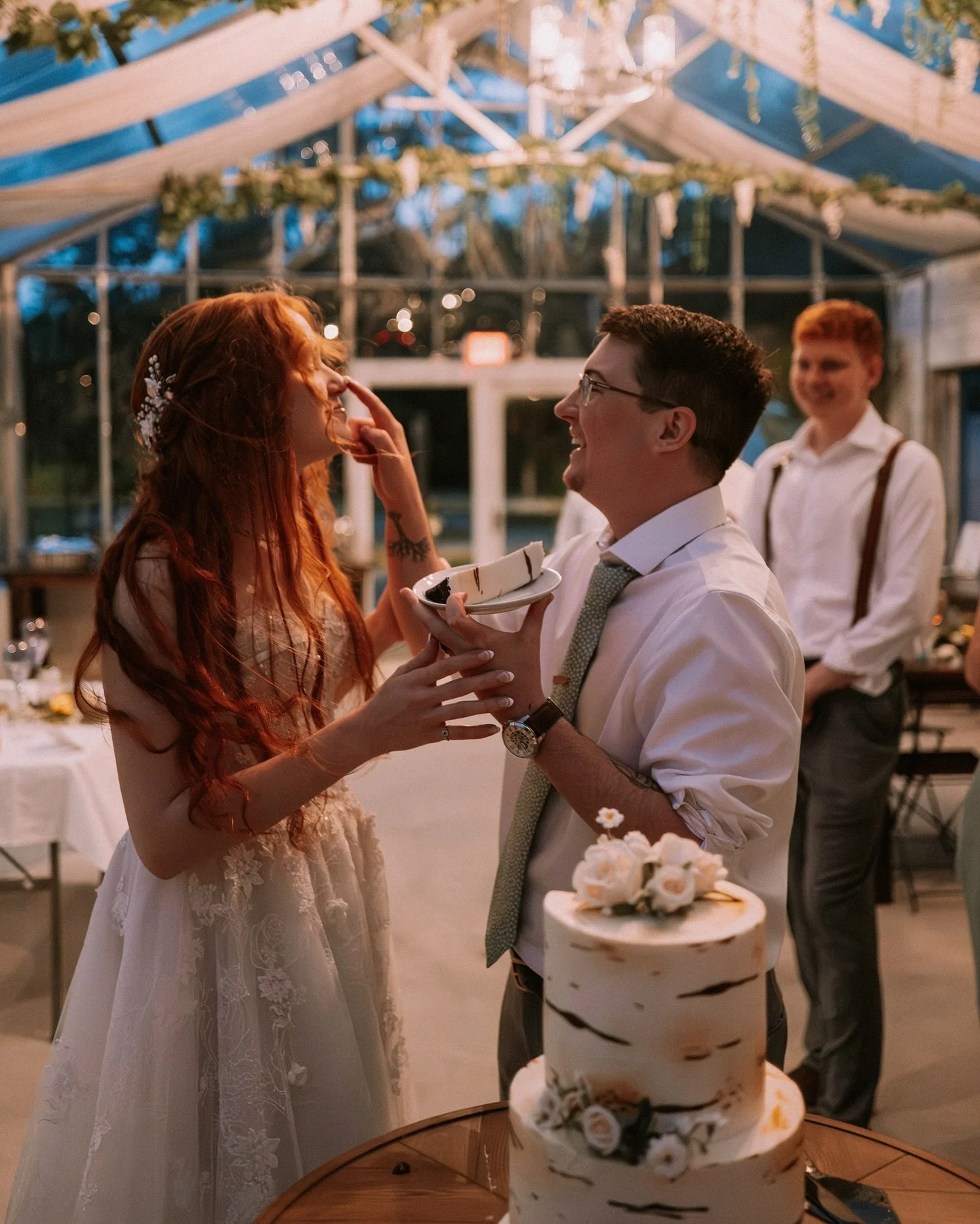A bride and groom feeding each other cake at a wedding reception inside a decorated tent with string lights.