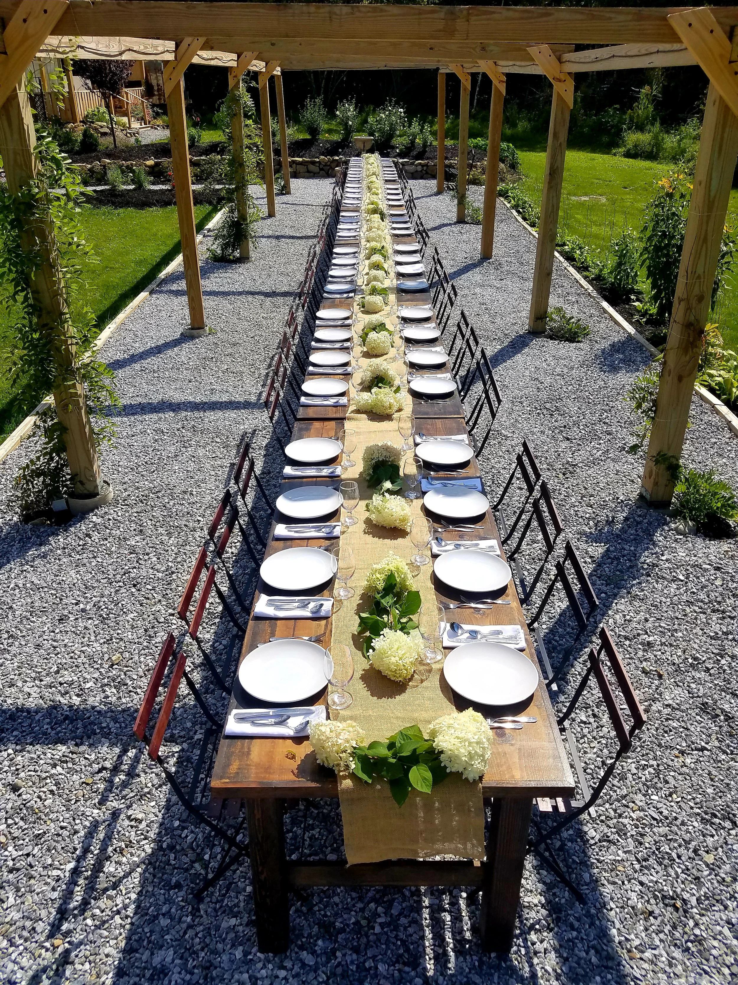 An outdoor dining setup with a long wooden table decorated with white flowers and greenery, set under a wooden pergola in a garden.